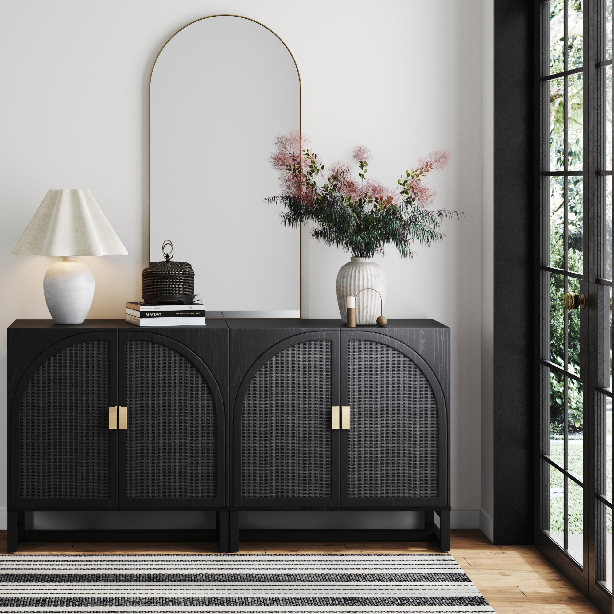 The Nathan James Arched Wooden TV Console Credenza Black Rattan (Set of 2) is topped with a lamp, decor, and pink flowers. A large arched mirror hangs above, natural light pours in, and a striped rug lies on the floor.