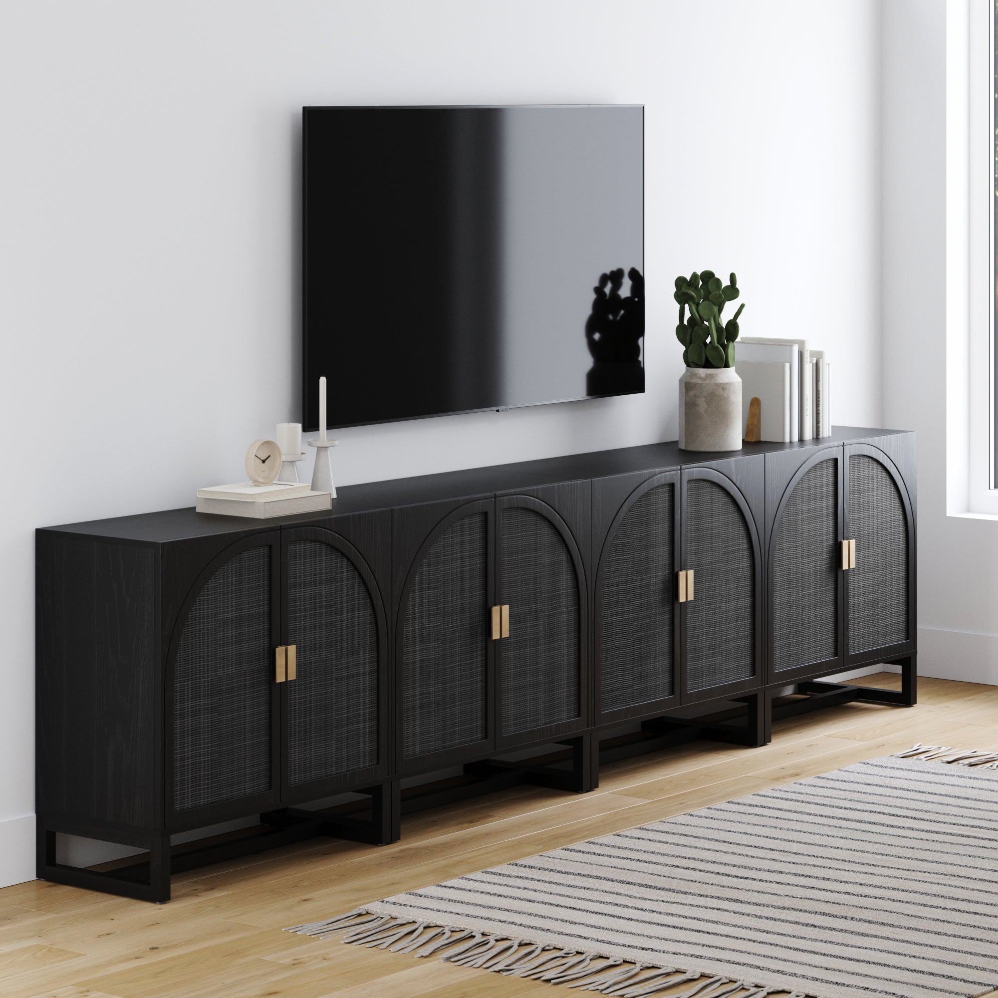 A modern living room features a large black TV mounted above the Nathan James Arched Wooden TV Console Credenza Black Rattan (Set of 4), decorated with books and accents. Light wood floors and a striped rug complete the inviting space.
