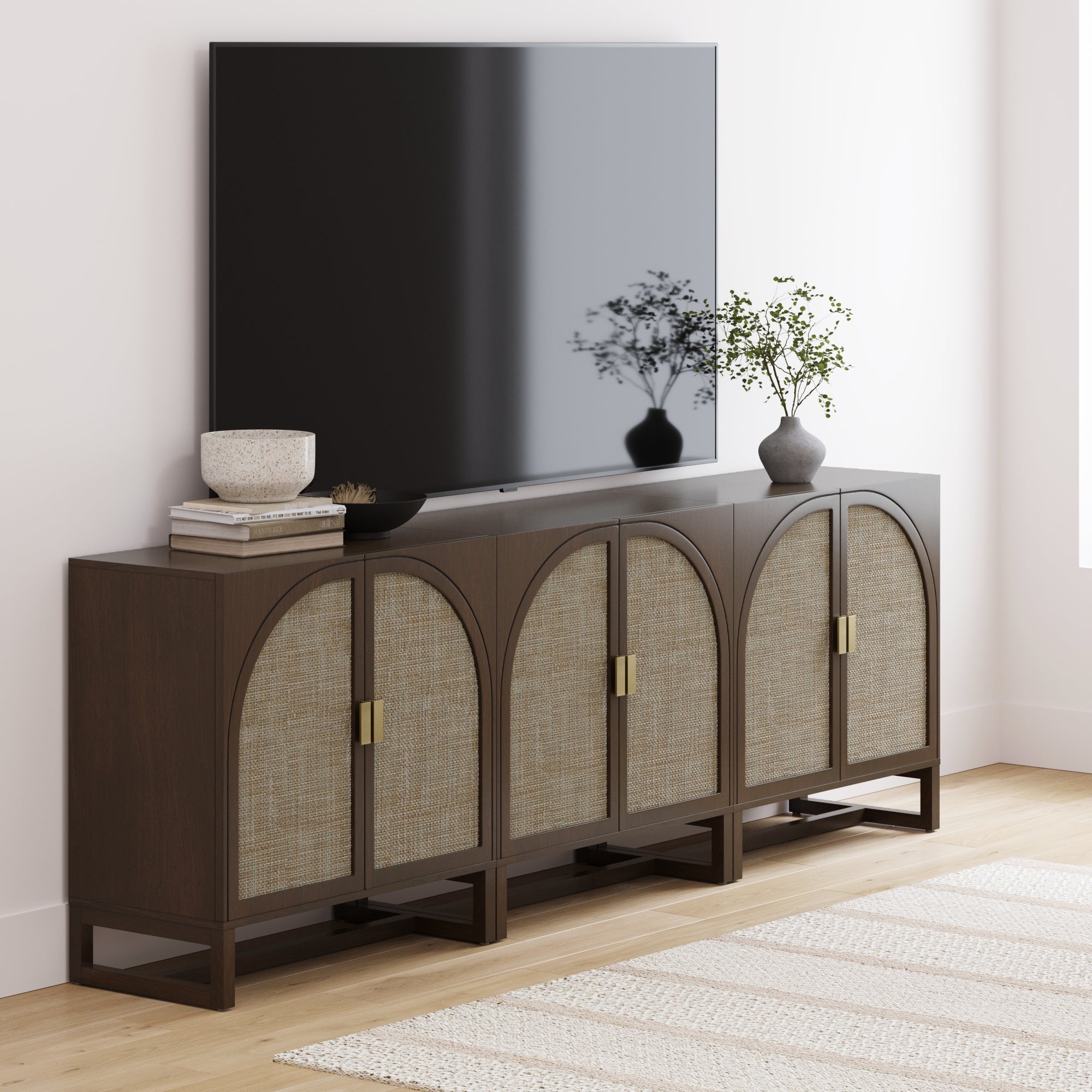 A flat-screen TV is mounted above the Nathan James Boho Arched TV Console Credenza in Dark Acacia (Set of 3). Decorative bowls and potted plants are displayed on top, with a light rug laid out over the wooden floor.