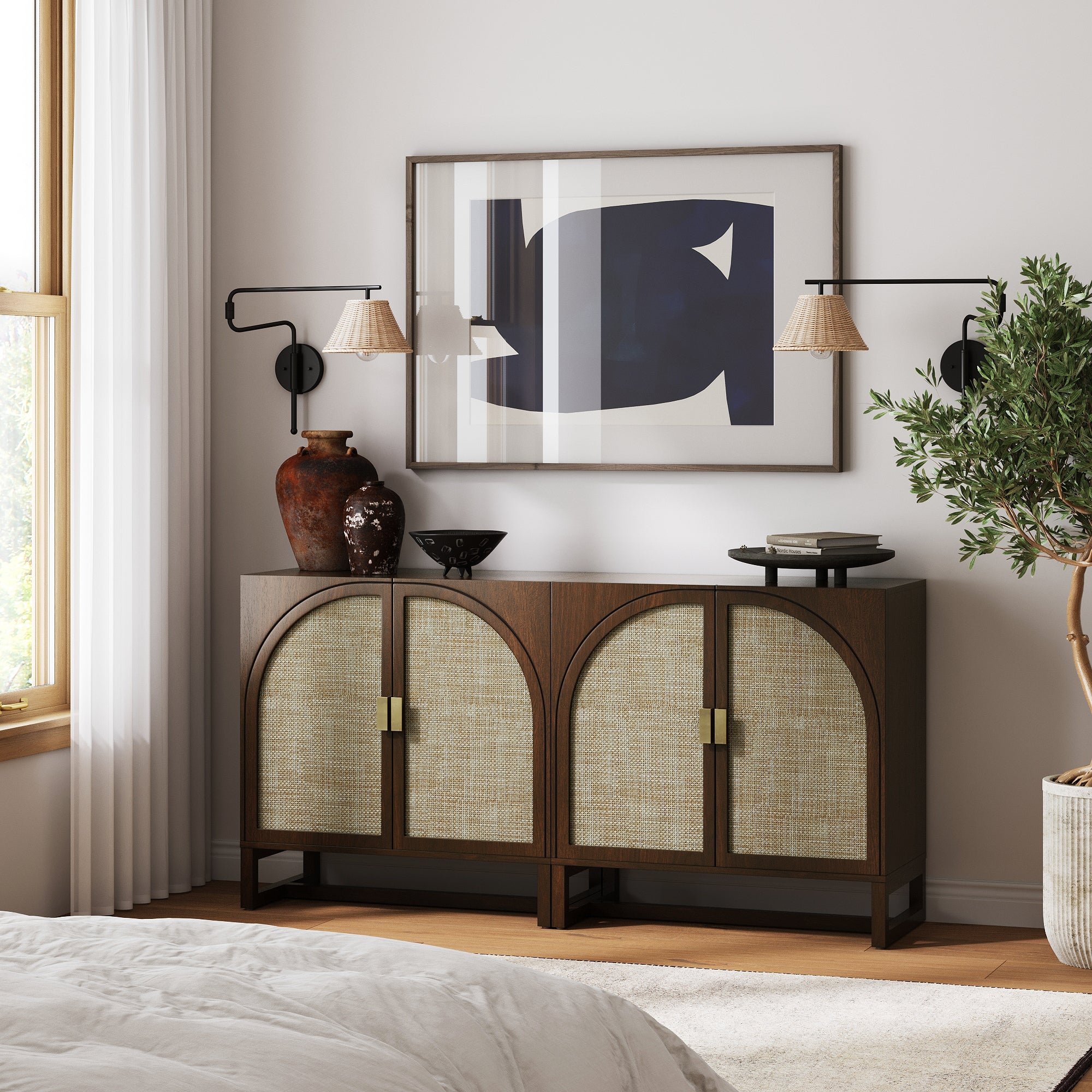 A stylish bedroom showcases the Nathan James Boho Rattan Arched Door Storage Cabinet in dark acacia, paired with ceramic vases, a black bowl, abstract art above, wall lamps on each side, and a leafy plant by the sunlit window.