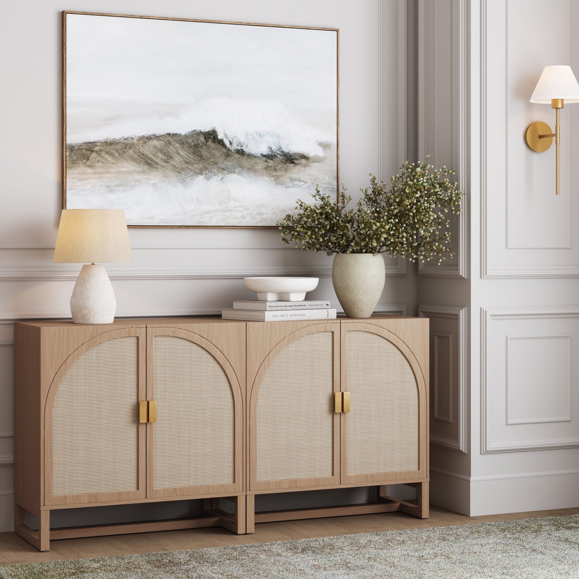 A light wood sideboard with curved cabinet doors is topped with a table lamp, decorative bowl, and a large vase of greenery. Above it hangs a framed abstract painting of a wave. The wall is white with elegant paneling.