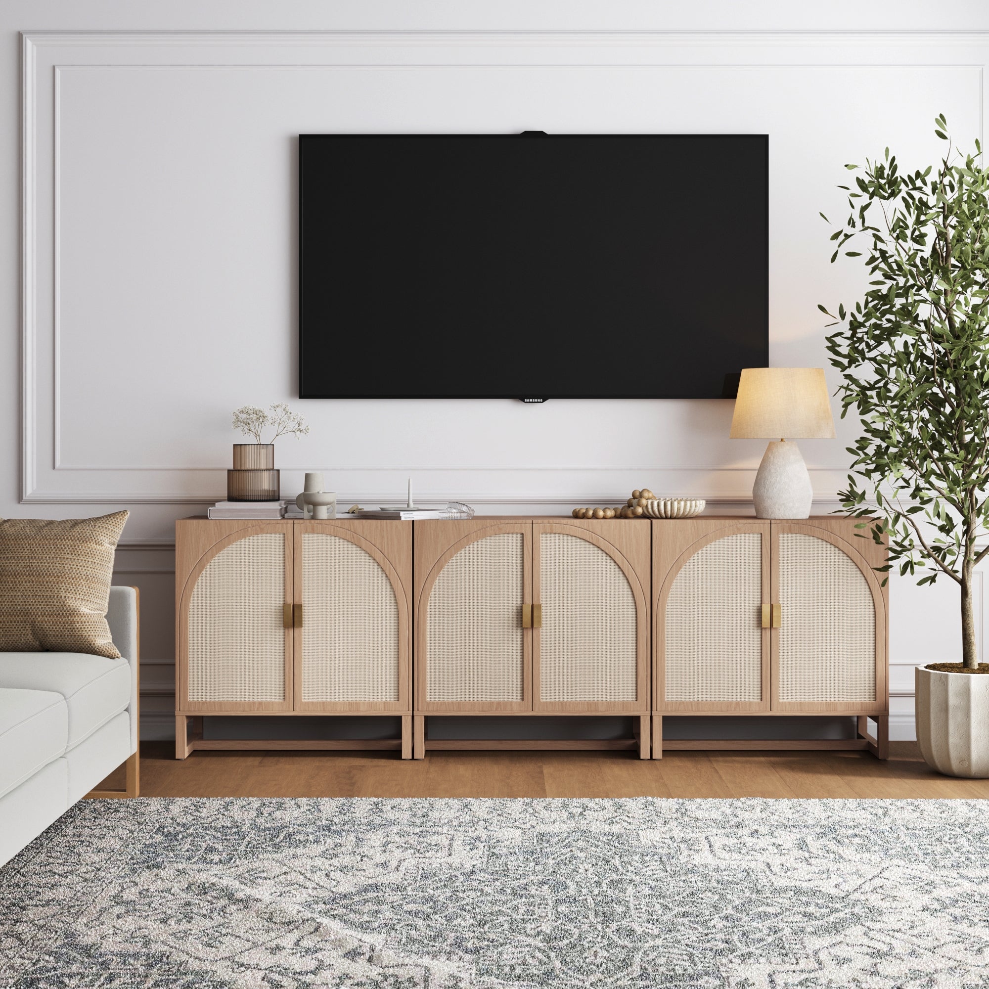 A modern living room features a large wall-mounted TV above the Nathan James Boho Arched TV Console Credenza Raw Oak (Set of 3), a lamp, decor, a potted plant, a patterned rug, and part of a white sofa.
