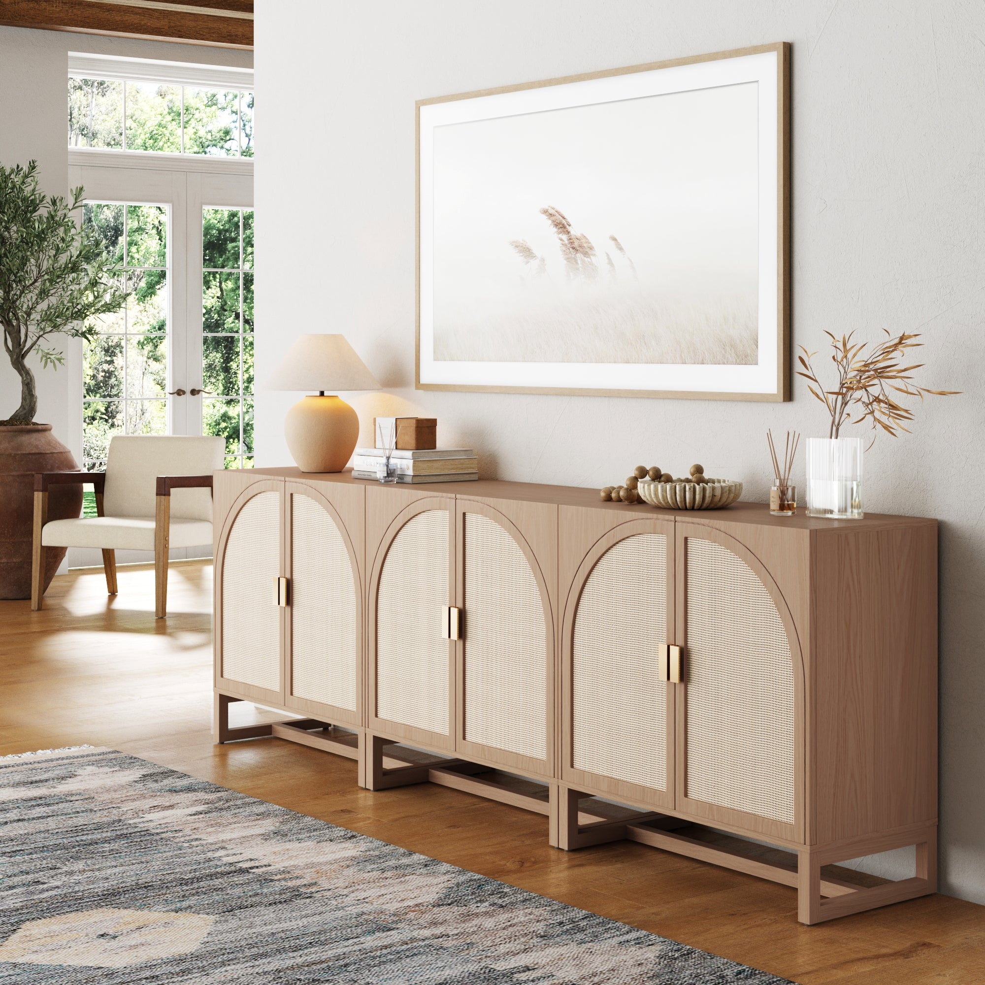 A modern living room showcases the Nathan James Boho Arched TV Console Credenza Raw Oak (Set of 3) with arched doors, topped with a lamp, books, and decor. A large framed photo hangs above, sunlight from glass doors brightening the neutral palette.