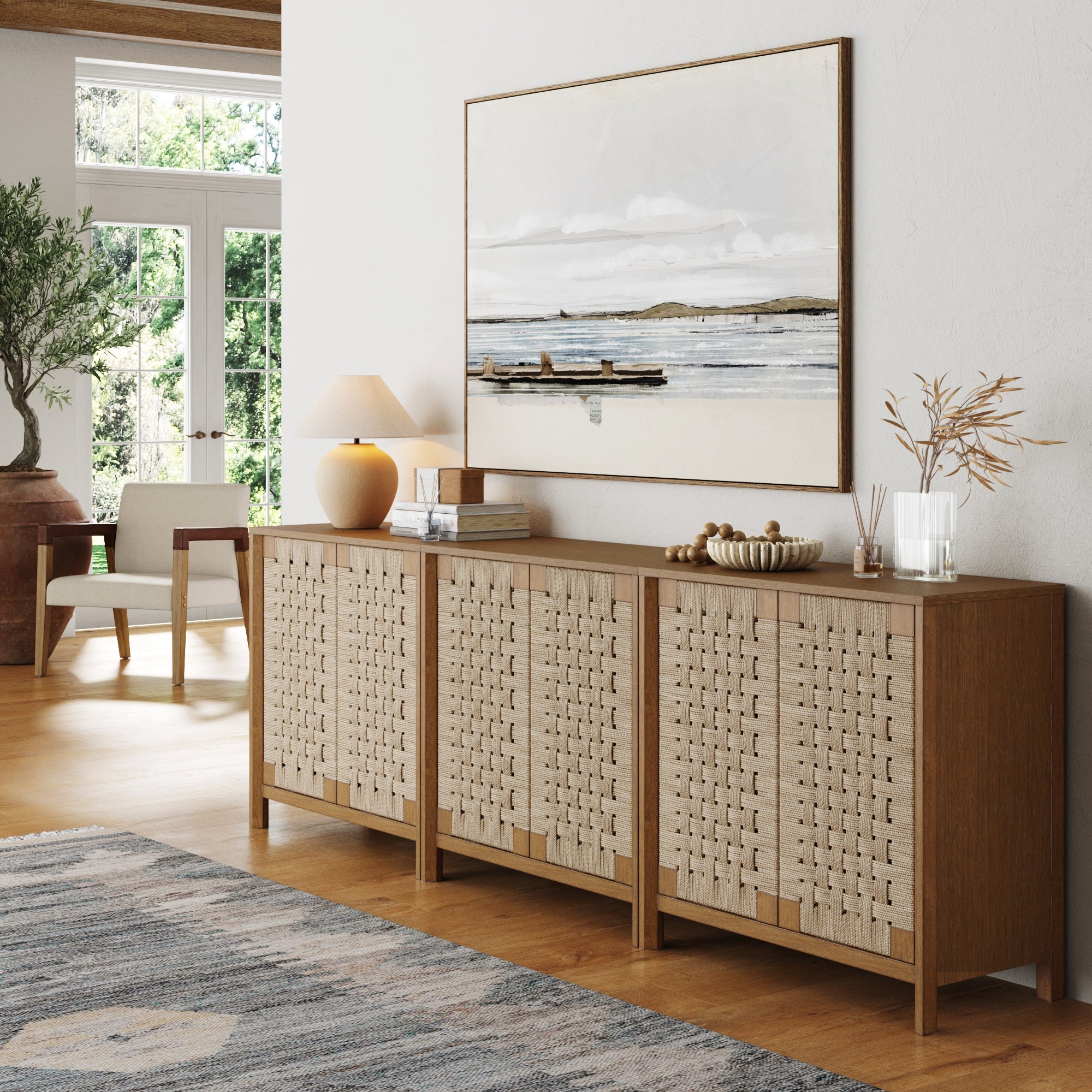 A modern living room with light wood floors and the Nathan James Boho Woven Seagrass TV Console Credenza (Set of 3), topped with a lamp, vases, books, plus a landscape painting, chair by the window, a corner plant, and streaming natural light.