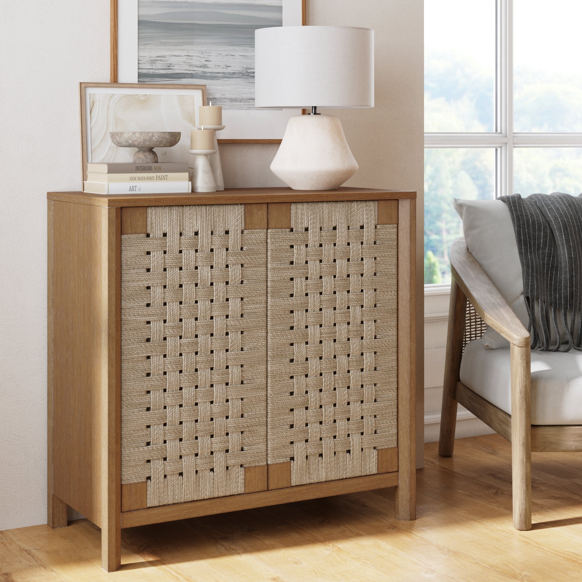 A wooden cabinet with woven beige doors is topped with a white table lamp, stacked books, and decorative objects. It stands beside a gray armchair by a large window in a bright, modern room.