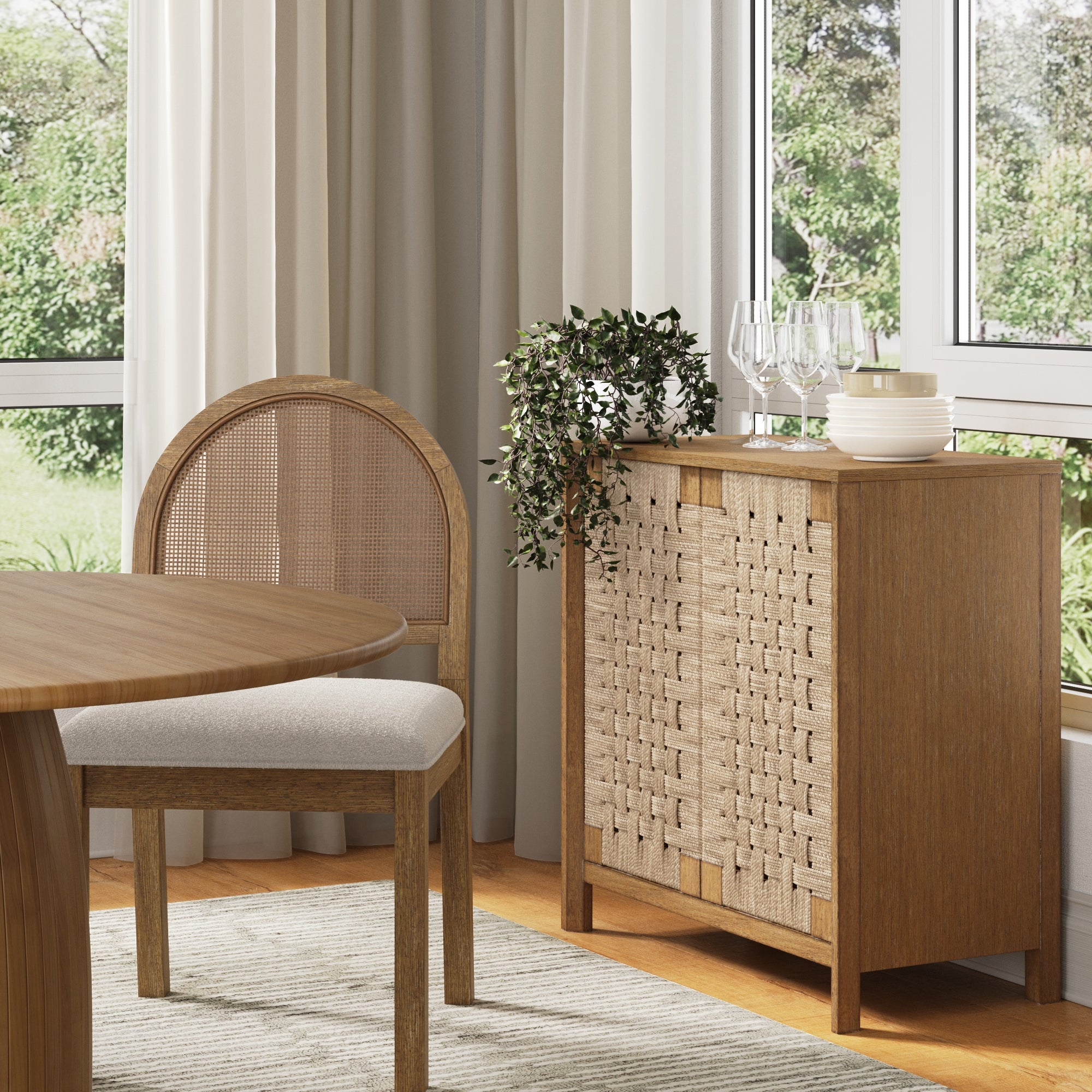 A wooden sideboard with a woven front stands next to a matching chair and round table in a bright, modern room with large windows, curtains, and a green view outside. Glasses and a plant sit on the sideboard.