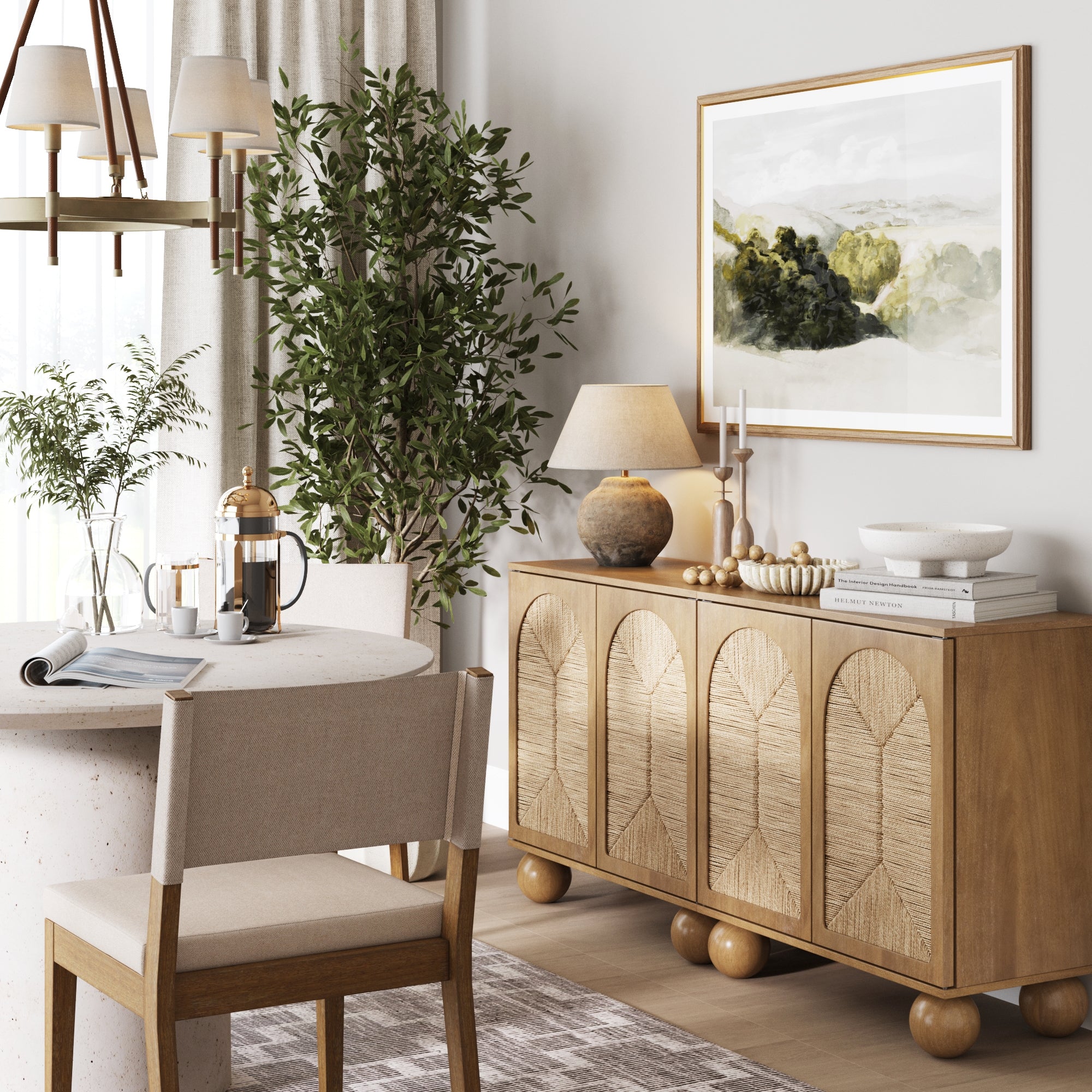 A cozy dining area features a round table, upholstered chair, and coffee setup. The Nathan James Seagrass Arched TV Console Credenza (Set of 2) in light brown adds boho style, while a large plant and framed art enhance the natural, neutral-toned ambiance.