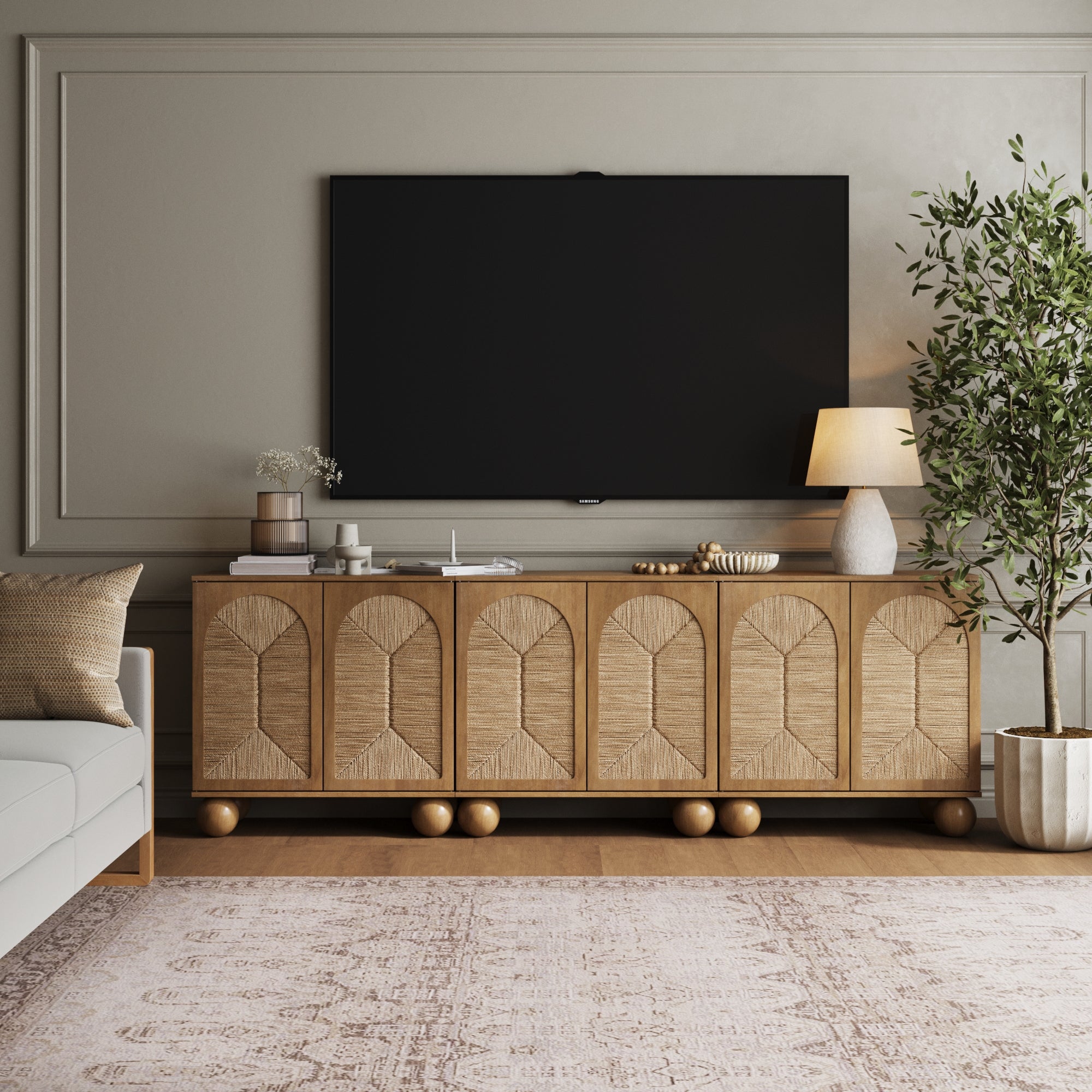 A modern living room features a large TV above the Nathan James Seagrass Arched TV Console Credenza Light Brown (Set of 3), with a potted plant, table lamp, decor, pale sofa, and patterned rug adding warmth to the minimalist, neutral-toned space.