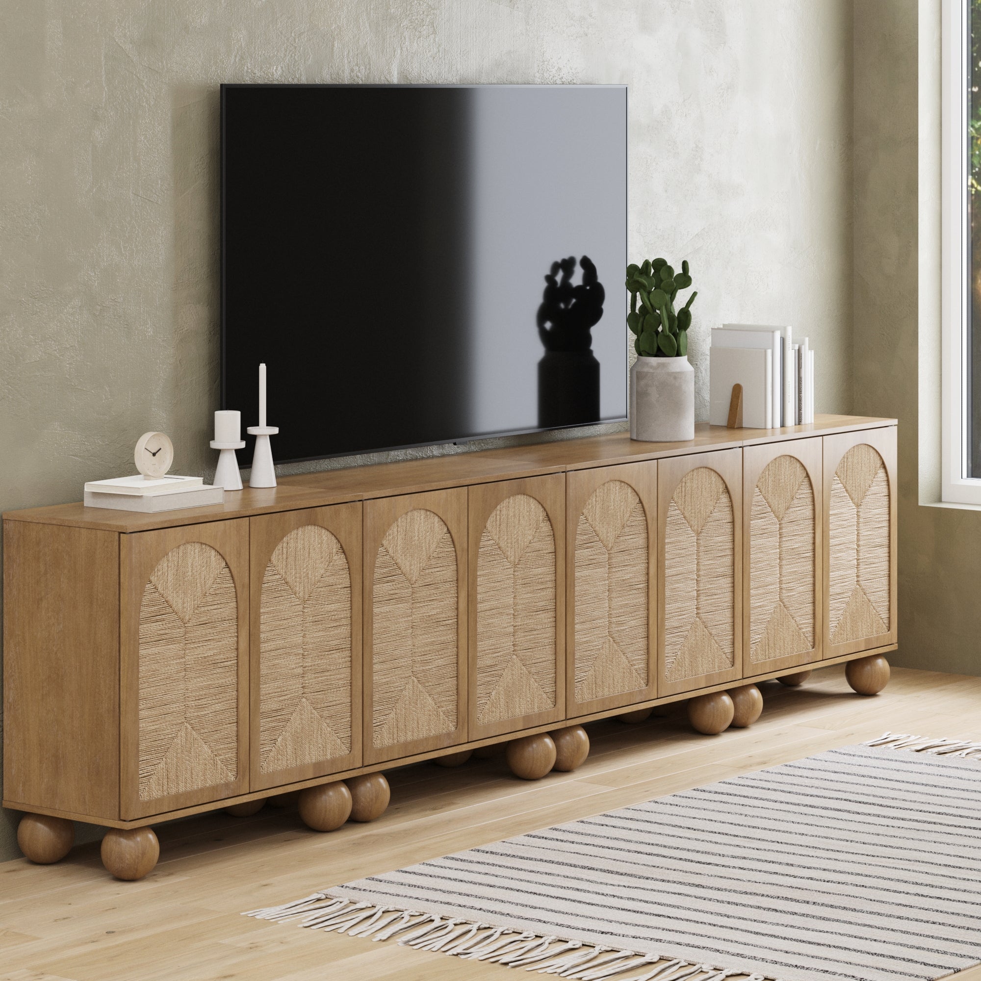 The Nathan James Large Seagrass Arched TV Console Credenza (Set of 4, Light Brown) displays a flat-screen TV, candles, a clock, a plant, and books. A striped rug and wooden flooring accentuate the minimalist living room style.
