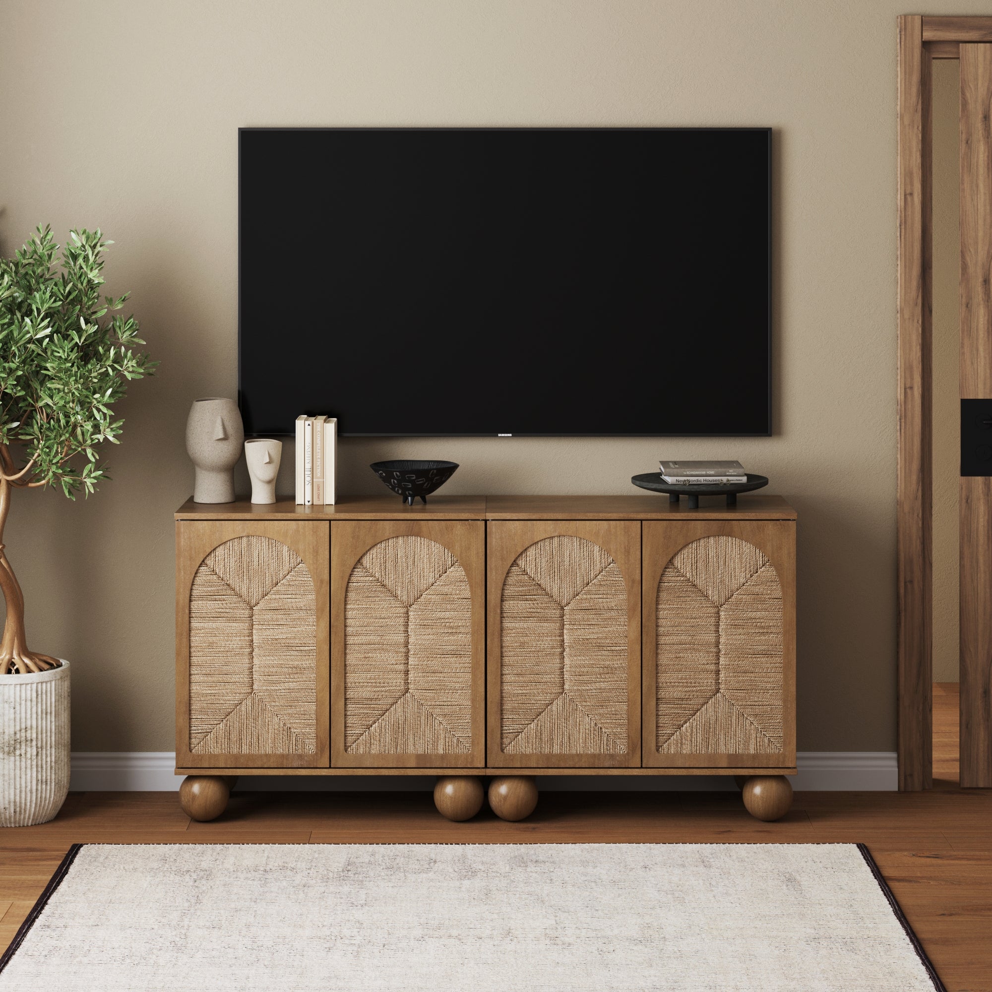 A modern living room features a large flat-screen TV mounted above the Nathan James Seagrass Arched Storage Cabinet in light brown. Decorative objects and books adorn the cabinet, with a potted plant standing to the left.