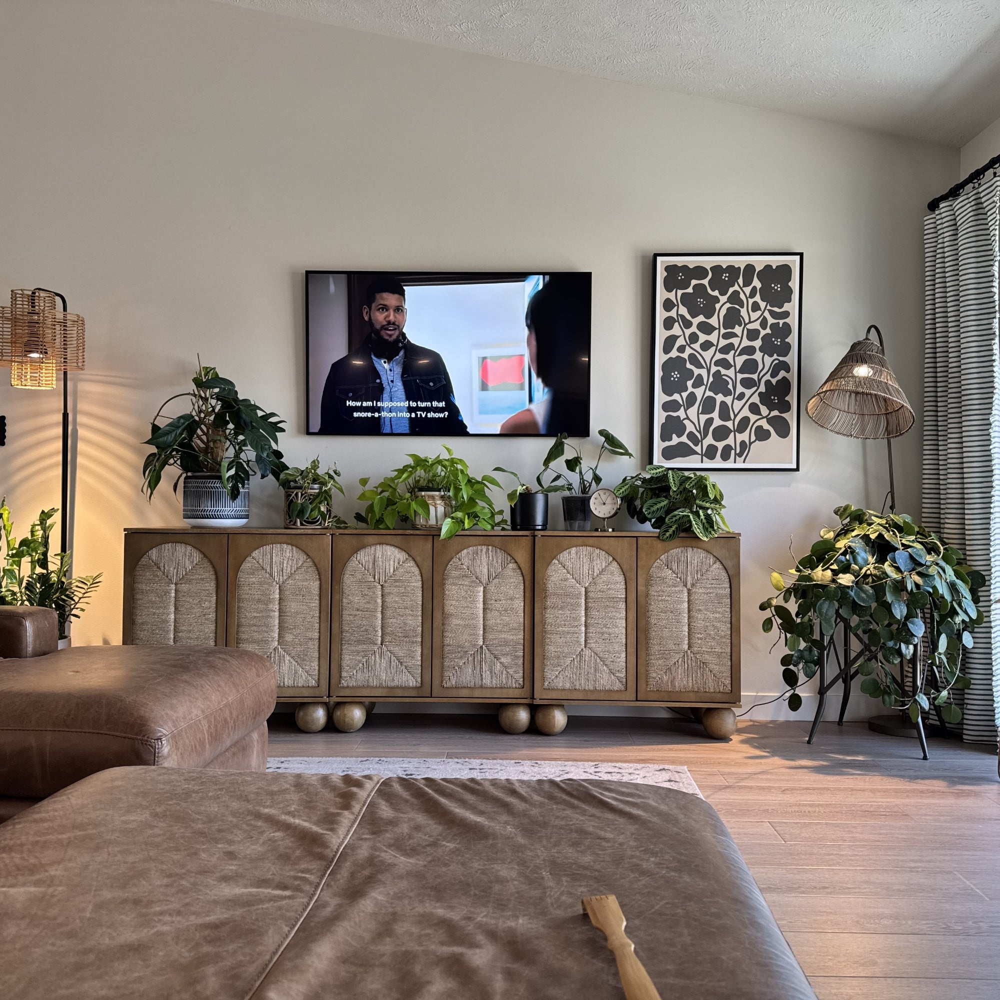 A cozy living room with a TV on the wall showing a man’s face, surrounded by green houseplants on a sideboard, a brown leather sofa, a black floor lamp, framed wall art, and light streaming through window blinds.