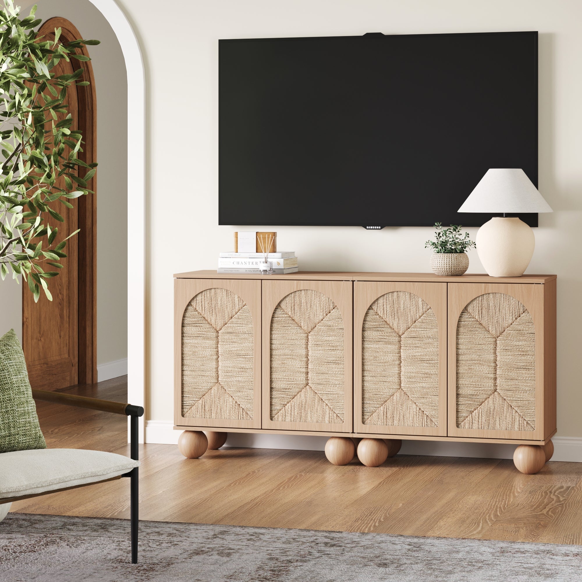 A modern living room features the Nathan James Seagrass Arched TV Console Credenza in Raw Oak (Set of 2) under a wall-mounted TV. The textured cabinets display decorative objects, a white lamp, and are accented by nearby greenery and part of a chair.