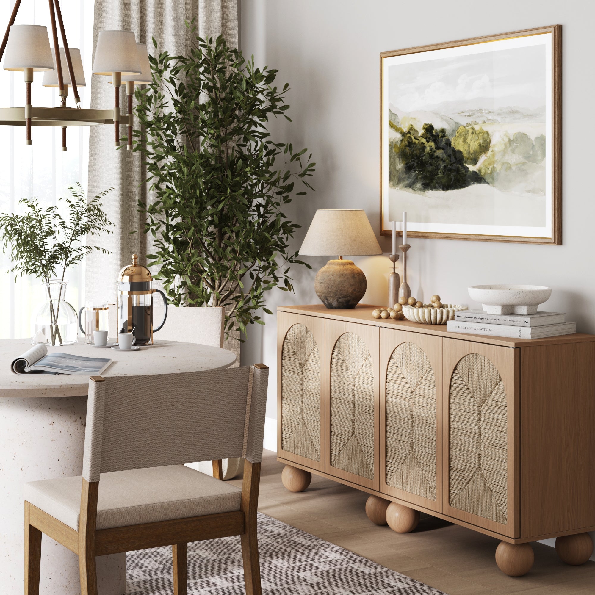 A cozy dining area with a round table, beige chair, and coffee set sits beside two Nathan James Seagrass Arched TV Console Credenzas in Raw Oak, styled with decor, a landscape painting, lamp on top, and lush greenery nearby.