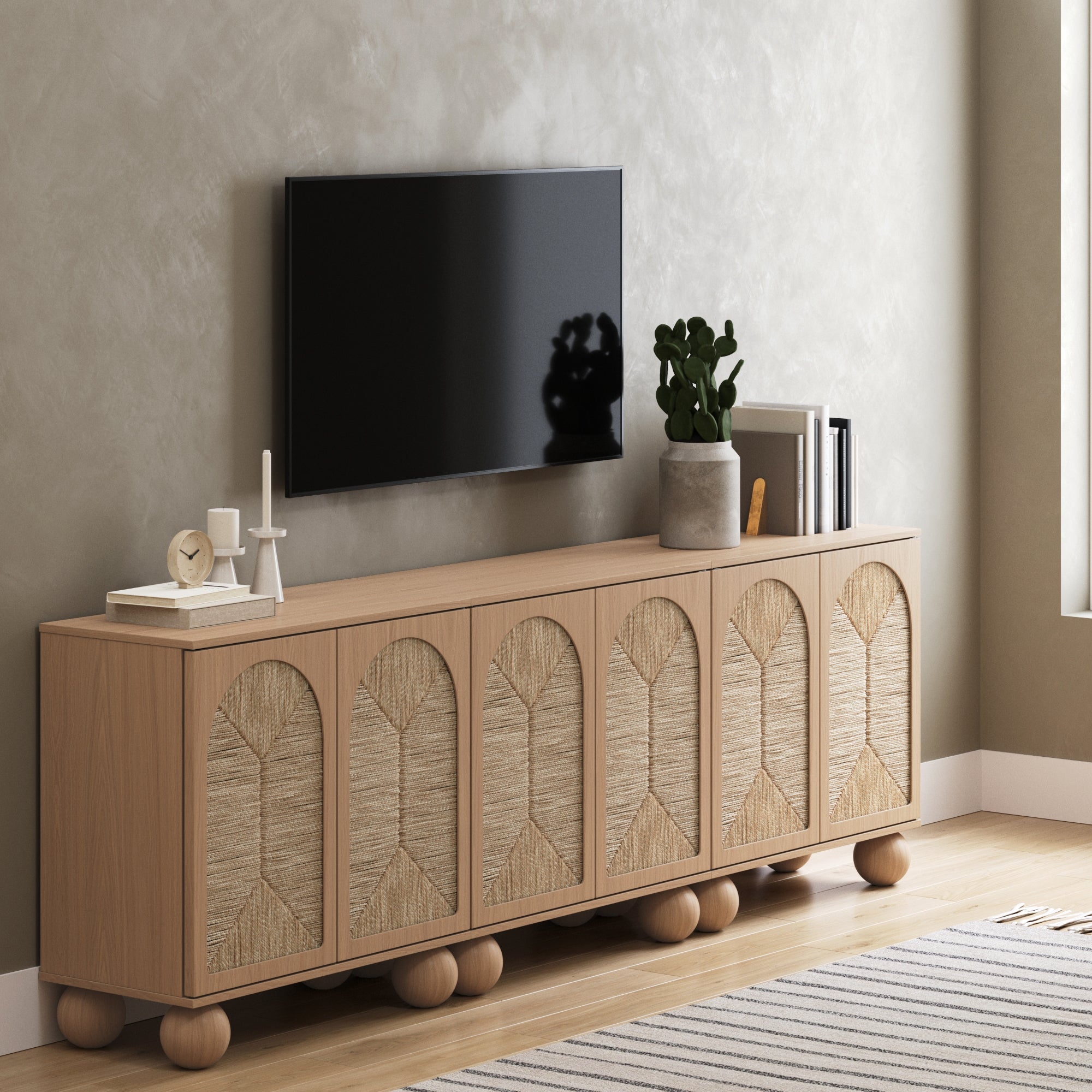 A modern living room with a wooden sideboard featuring woven panel doors, decorative items including candles, books, and a plant on top, and a flat-screen TV mounted on the light-colored wall above.