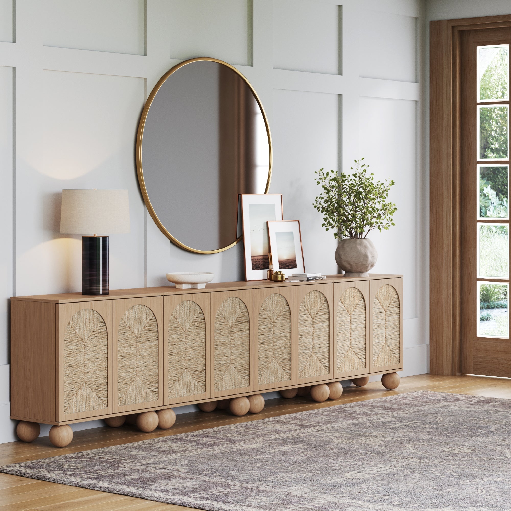 A modern living room features a wooden sideboard with decorative panels, a round mirror above it, a lamp, framed photos, and a plant. The space has wainscoted walls, a patterned rug, and natural light from a glass door.