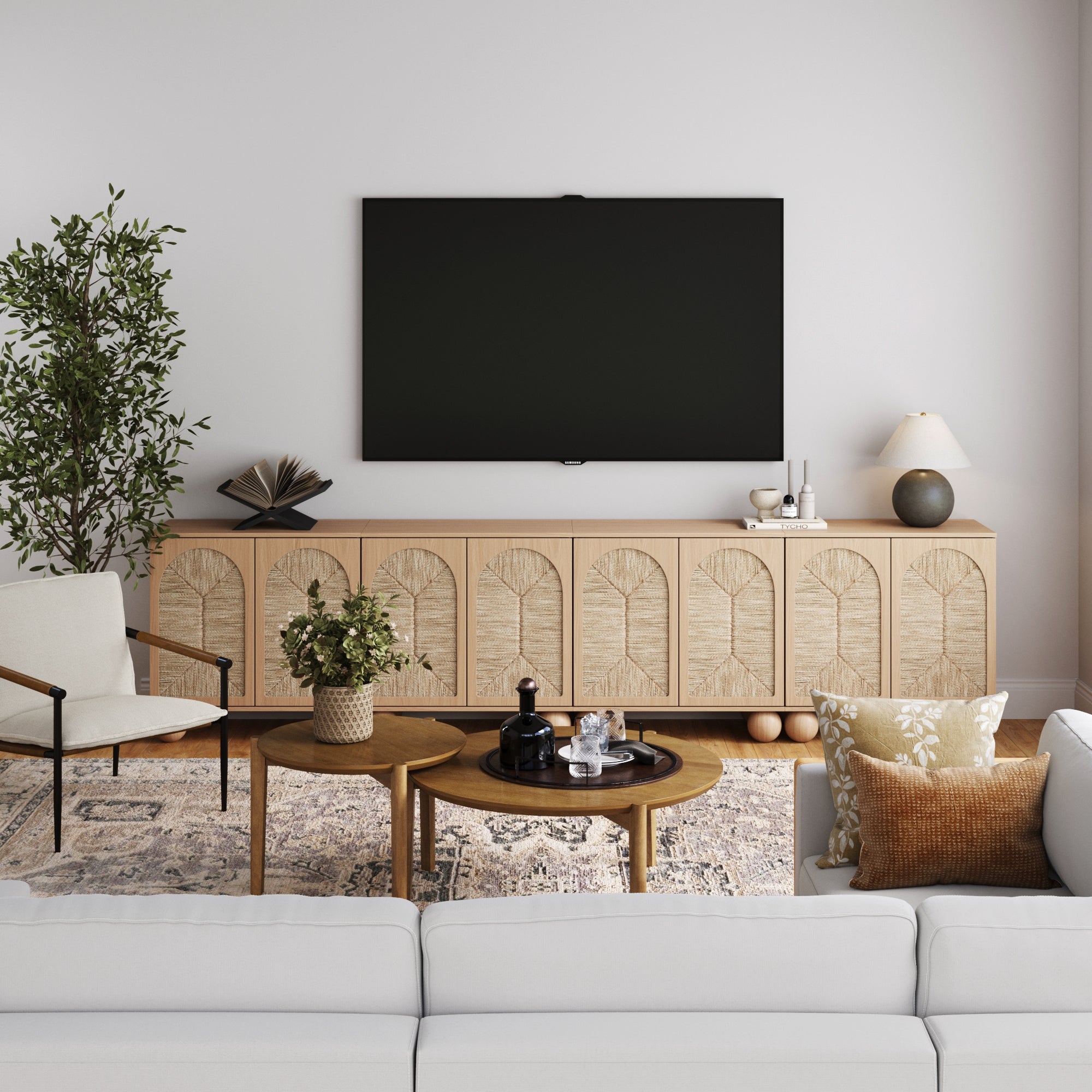 A modern living room features a light sofa, wooden coffee tables, the Nathan James Large Seagrass Arched TV Console Credenza in Raw Oak (Set of 4), plants, neutral decor, and patterned cushions for a cozy, stylish vibe.