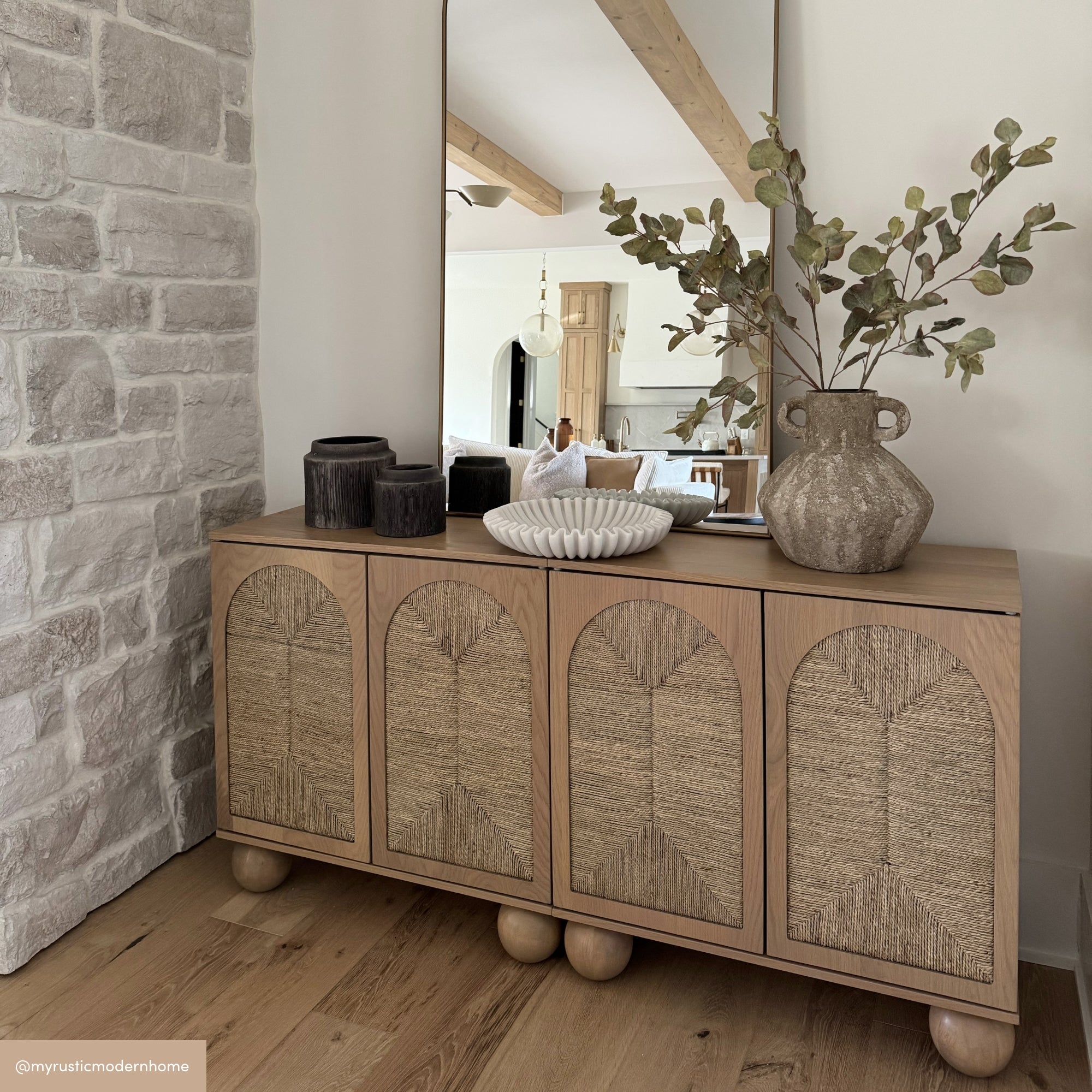 A modern rustic sideboard with four textured cabinet doors stands against a stone wall. On top are decorative items: two dark vases, a white sculptural dish, and a large jug with eucalyptus branches. A tall mirror reflects a cozy living room.