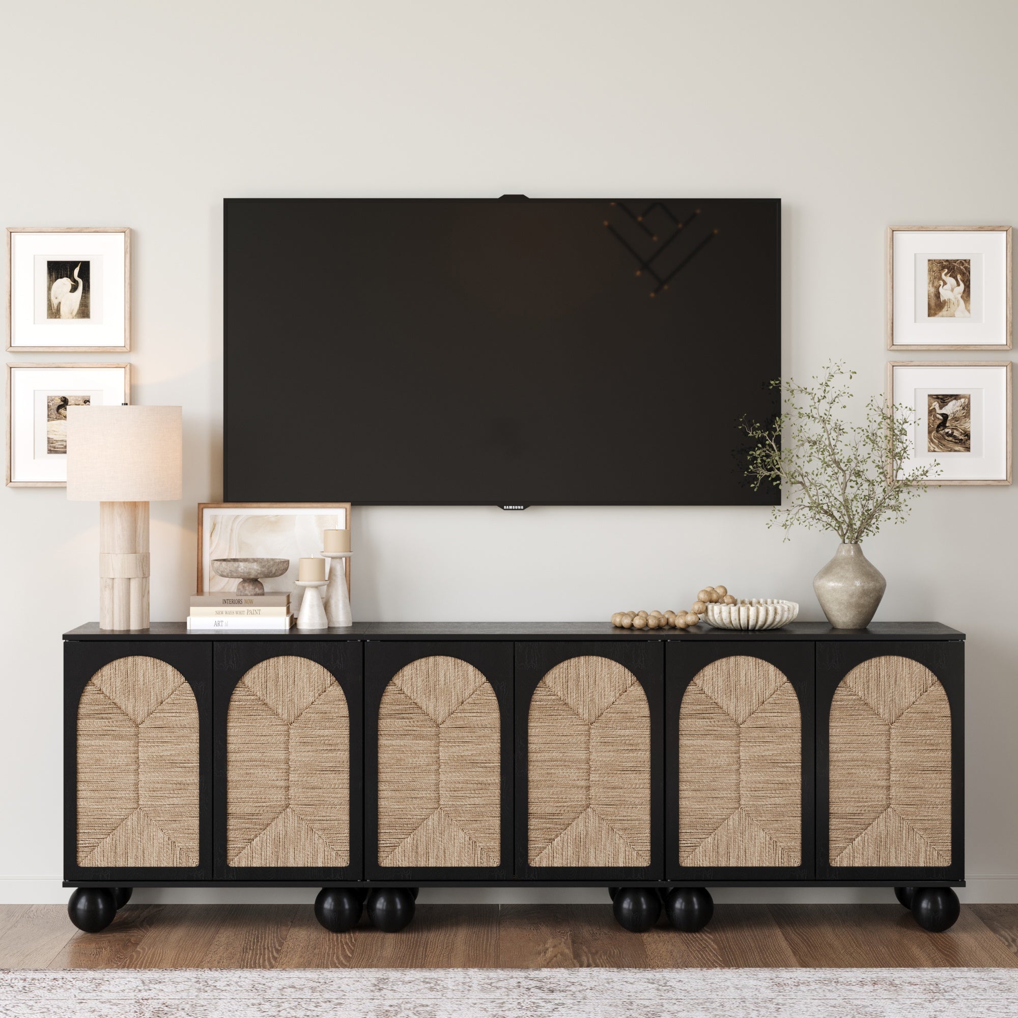 A modern living room features the Nathan James Seagrass Arched TV Console Credenza Black (Set of 3) with woven seagrass doors, a wall-mounted TV above, framed art, a lamp, stacked books, decor accents, and a vase with greenery on the console.