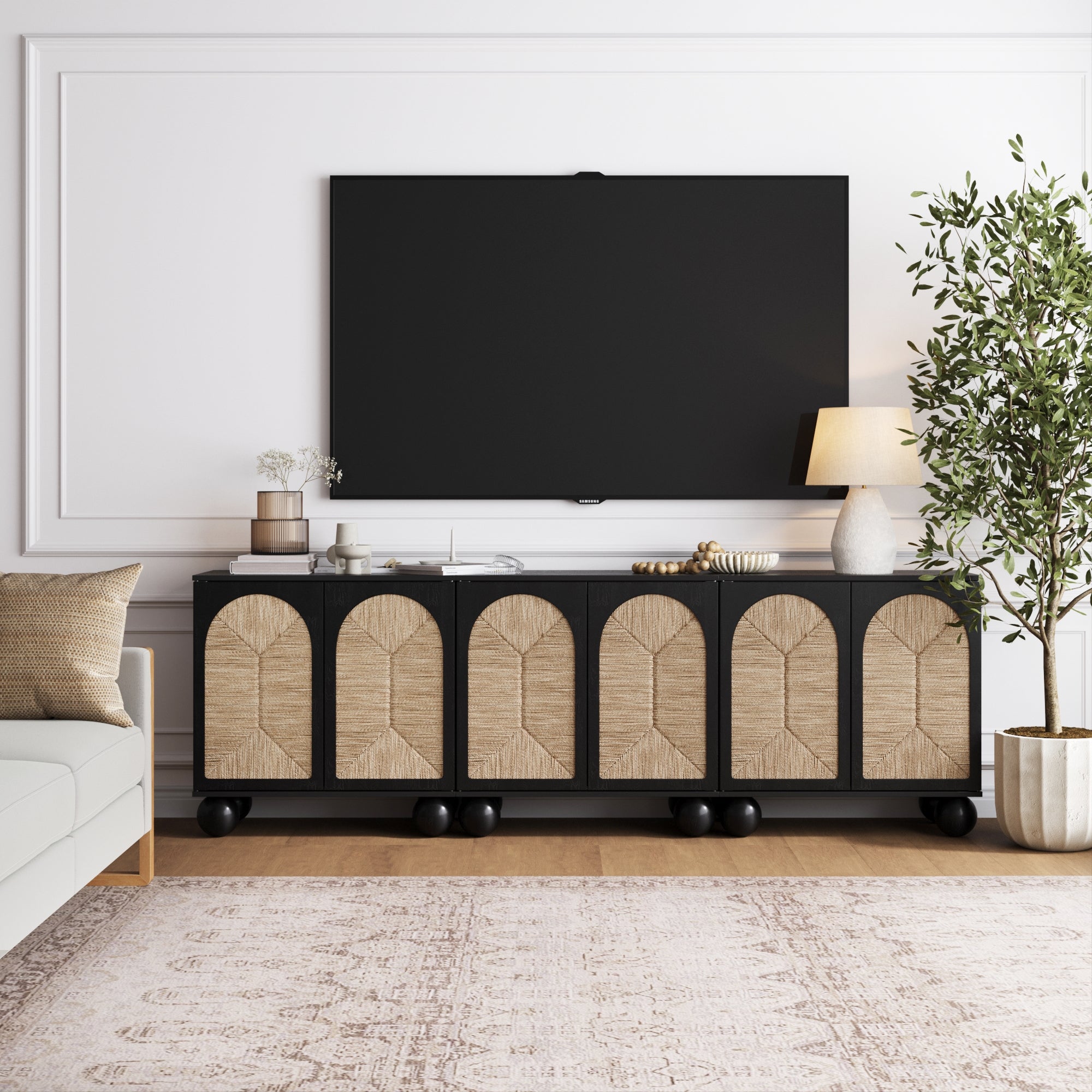 A modern living room features the Nathan James Seagrass Arched TV Console Credenza in black (set of 3) beneath a mounted TV, with decorative items, a beige sofa, potted plant, table lamp, and light rug for effortless boho elegance.