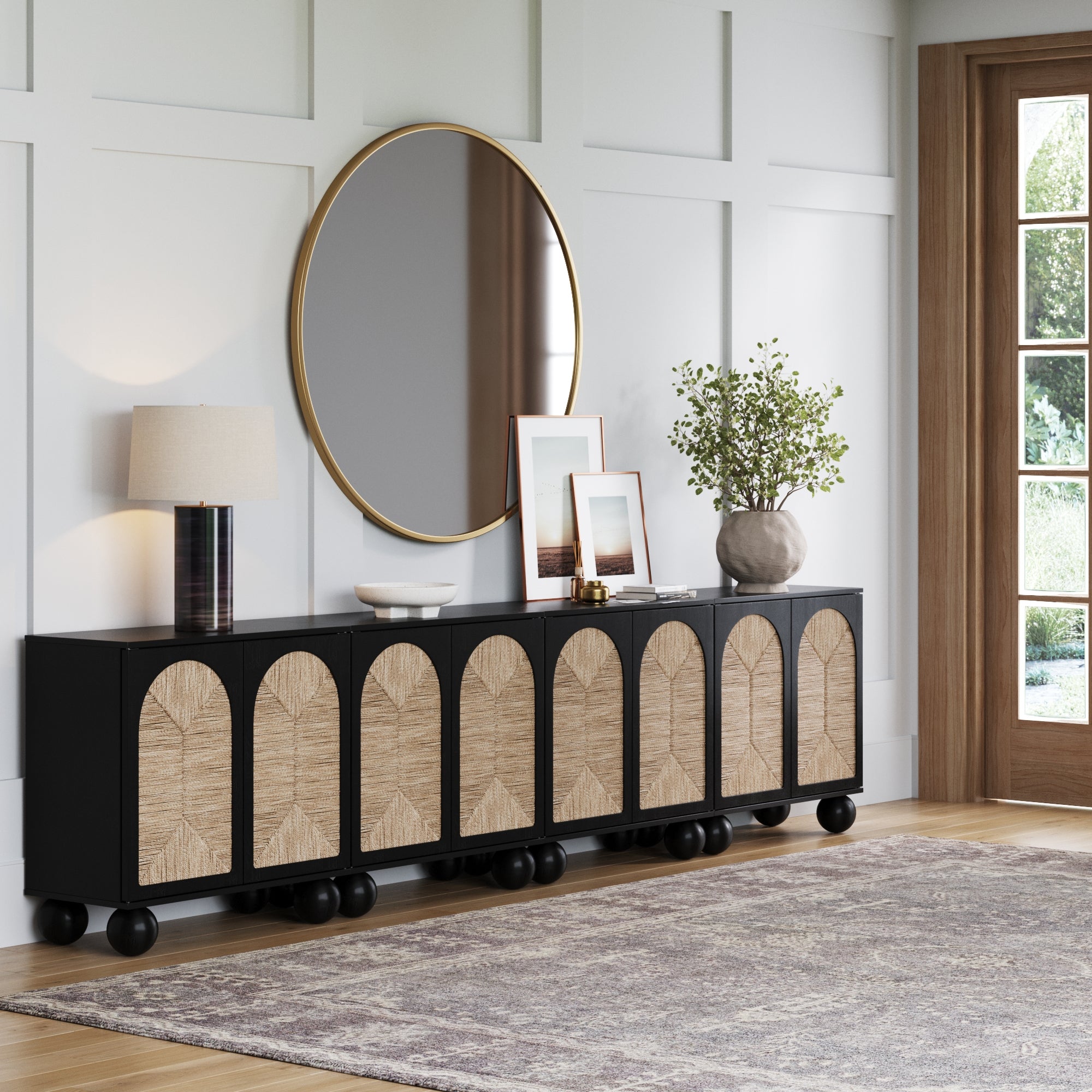 The Nathan James Large Seagrass Arched TV Console Credenza (Black, Set of 4) features arched wood panel doors and sits against a white paneled wall. A lamp, art, vase with greenery, and a large round mirror above add boho elegance.