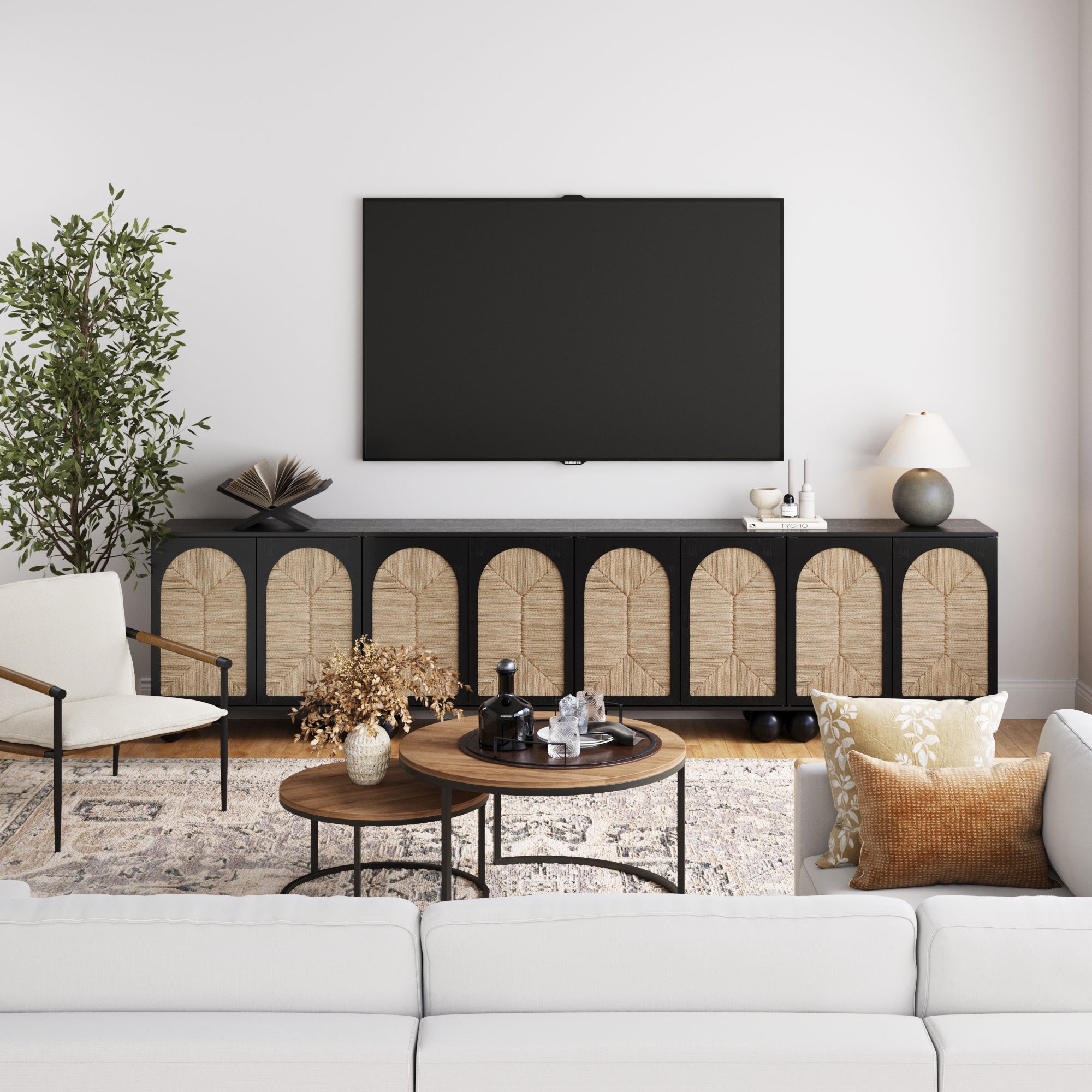 A modern living room features a white sofa, nested round wooden coffee tables, woven seagrass accents, a Nathan James Large Seagrass Arched TV Console Credenza Black (Set of 4), a large TV, tall plant, and neutral decor on a patterned rug.