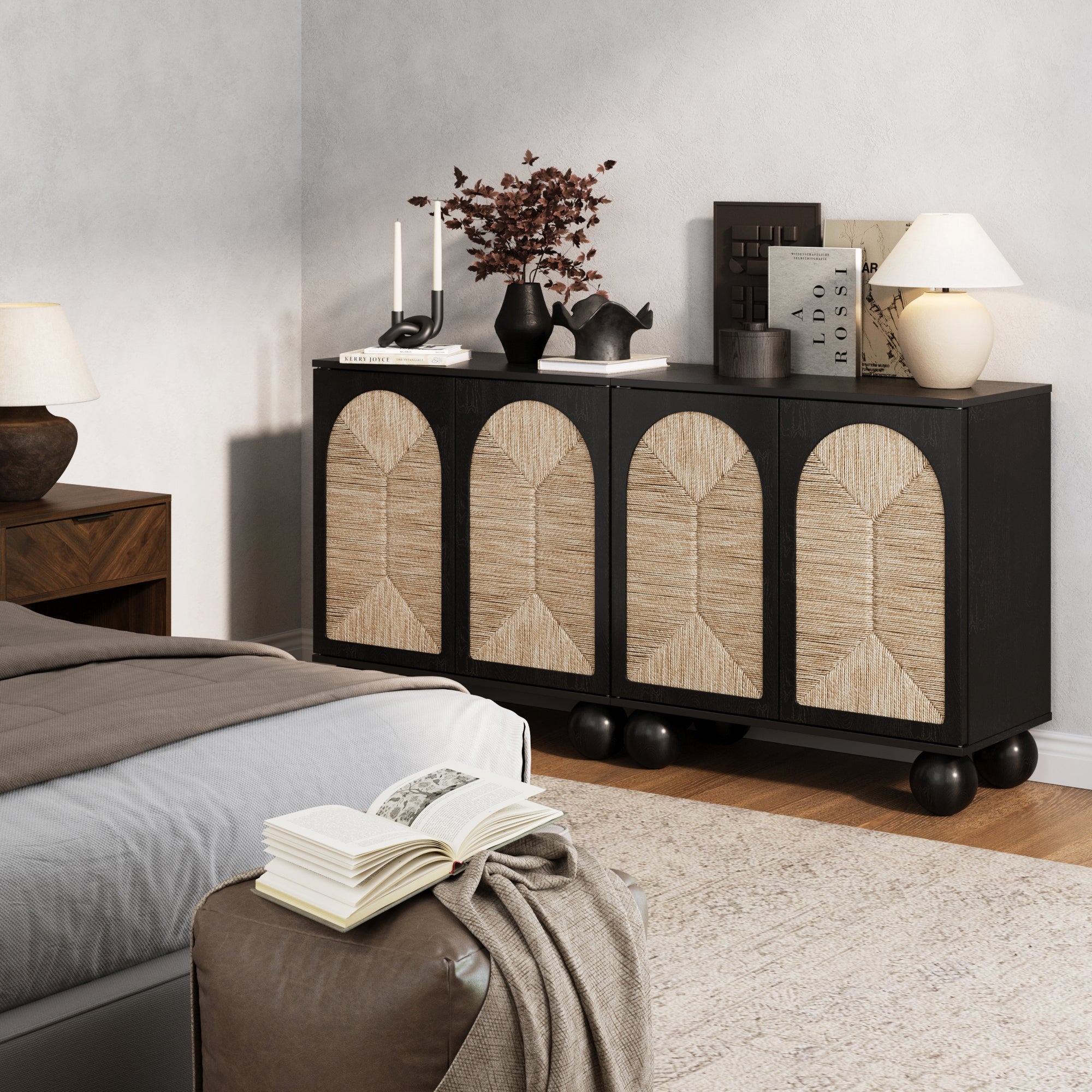 A stylish bedroom features the Nathan James Seagrass Arched Storage Cabinet in black, with rattan doors, topped with décor, books, lamps, and a vase of dried branches. An open book sits on a brown ottoman next to the bed.