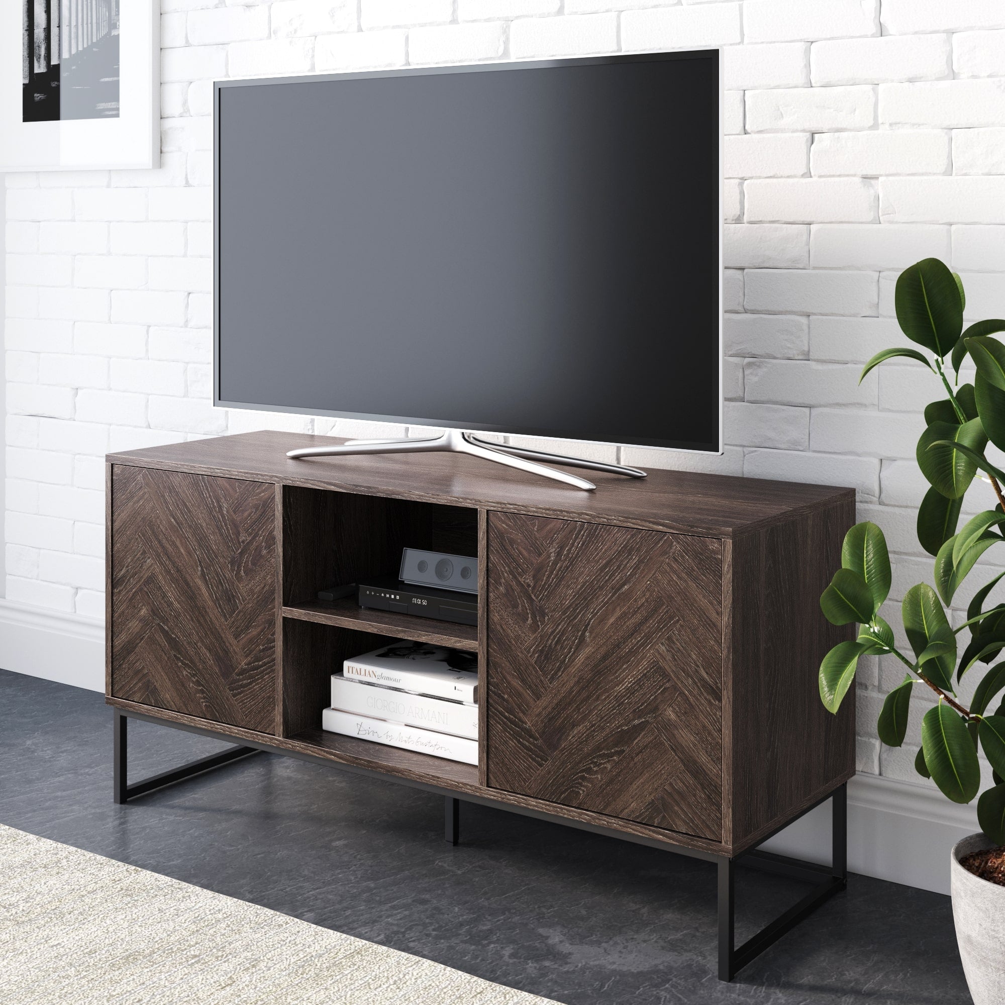A flat-screen TV sits on the Nathan James Wood Herringbone TV Cabinet in Light Golden Oak, its open shelves displaying books and electronics against a white brick wall, with a leafy green plant placed beside the stand.