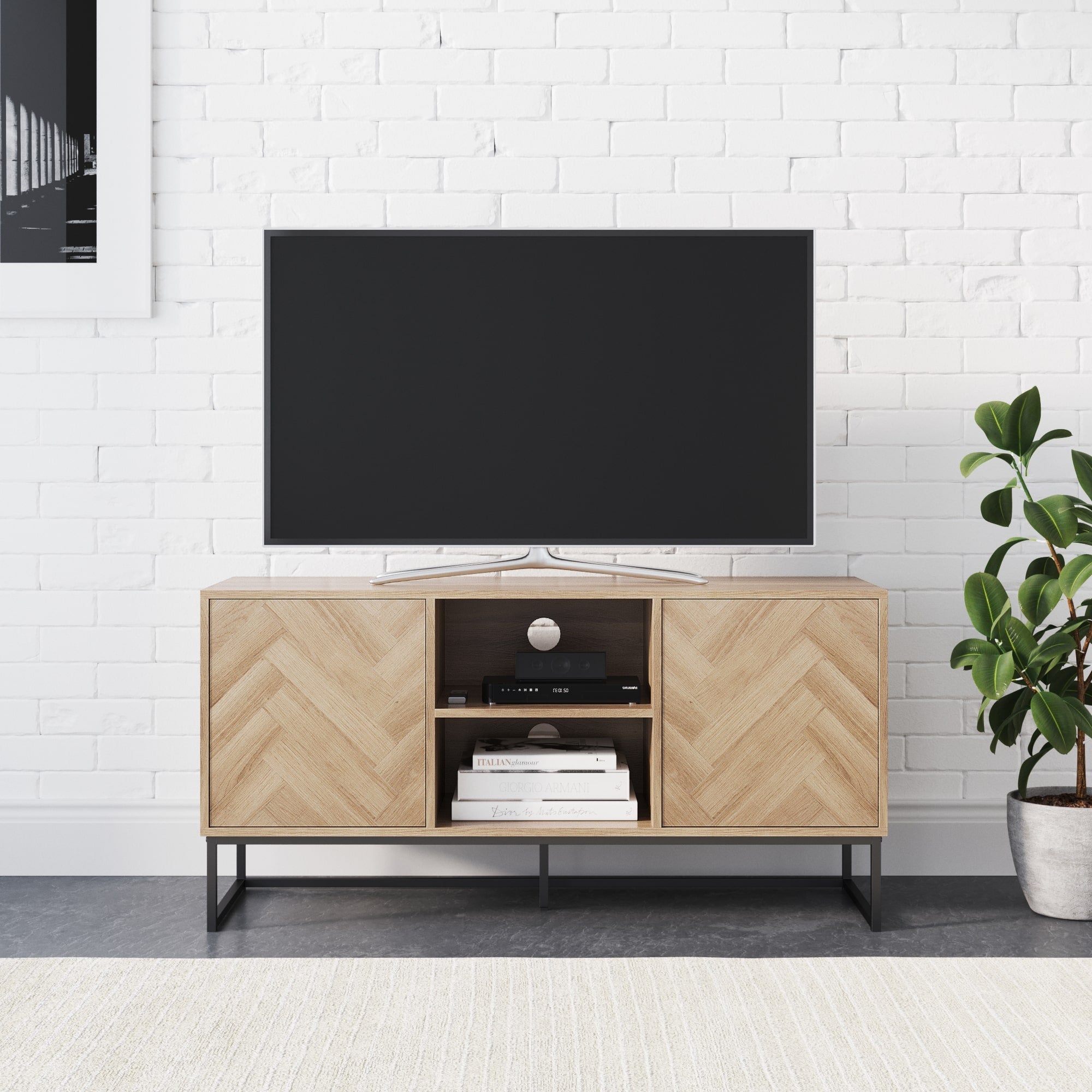 A flat-screen TV sits on a modern wooden TV stand with a chevron pattern, featuring open shelves with books and electronics. A green potted plant is on the right, against a white brick wall.