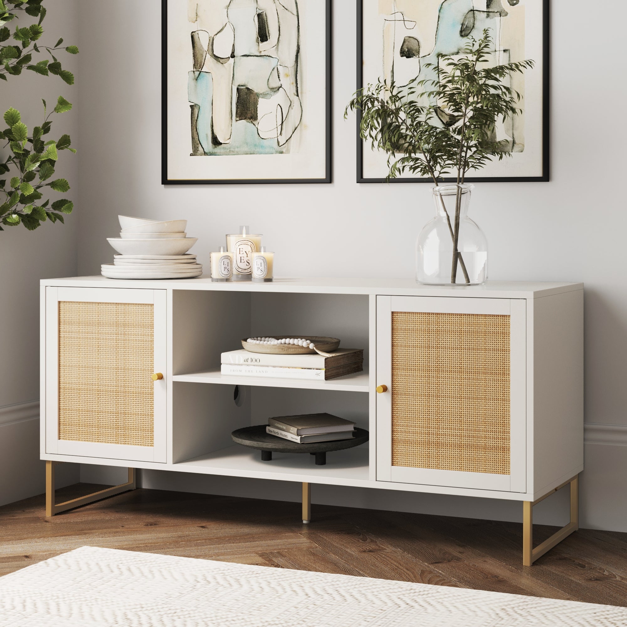 The Nathan James Rattan & Wood 2-Door TV Cabinet White features rattan doors, gold legs, and open shelves for decor, books, or plates. A glass vase with leafy branches tops it, with abstract art prints and a green plant in the background.