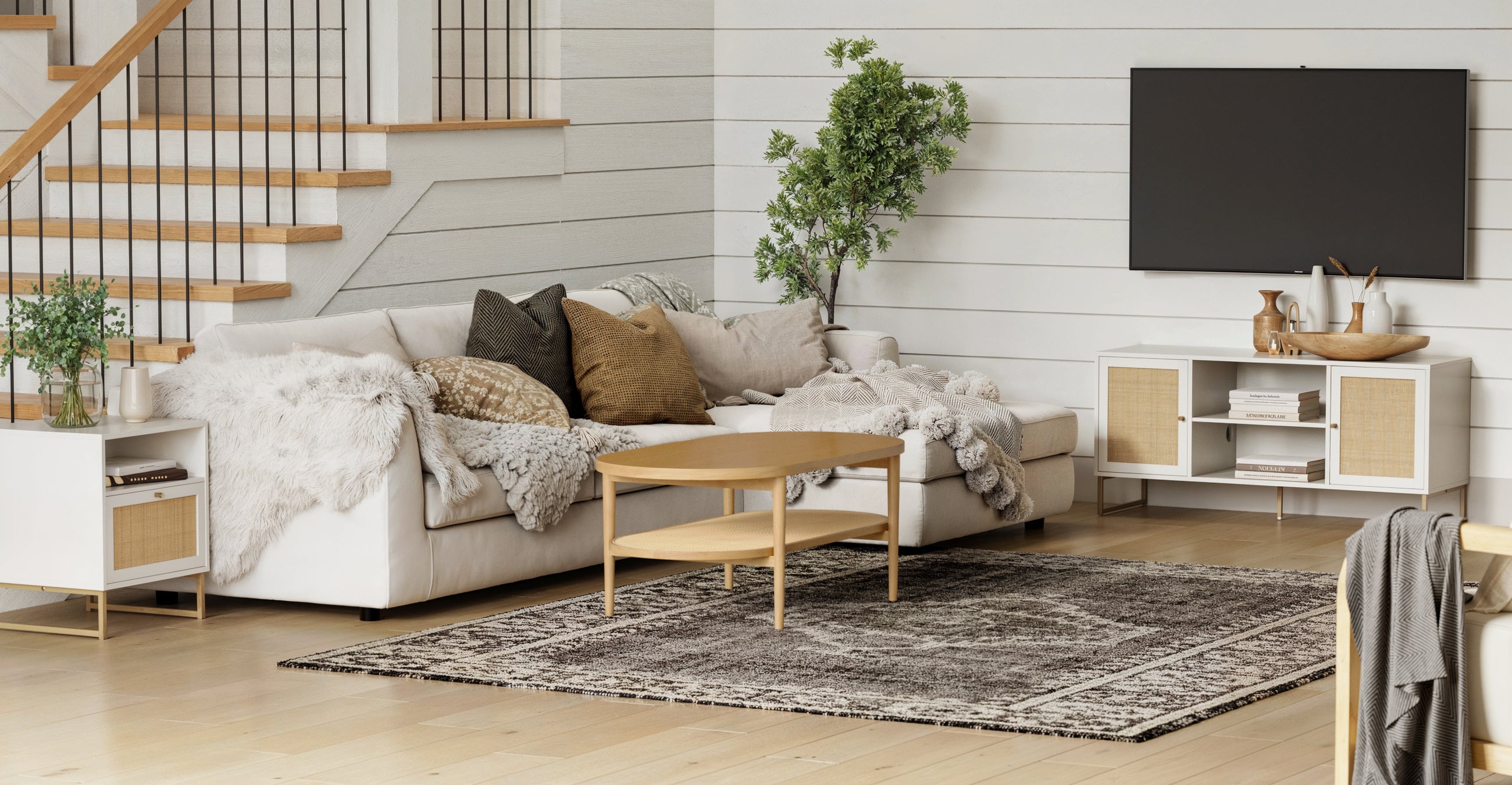 A cozy modern living room with a white sectional sofa, neutral pillows and blankets, a round wooden coffee table, a patterned rug, a potted plant, and a TV mounted on the wall next to a staircase.