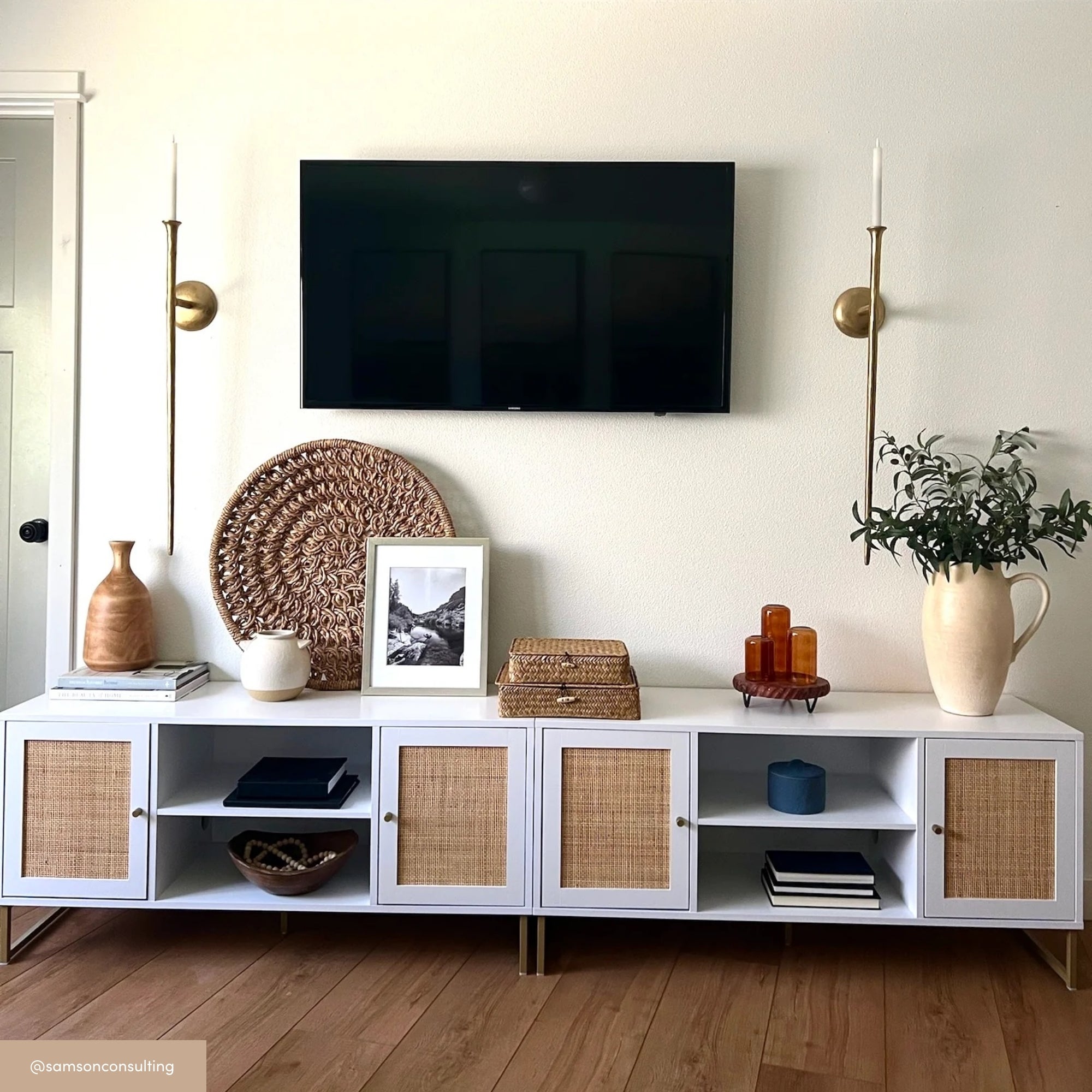 A modern white TV console with wicker door panels sits below a wall-mounted TV. Decorative items include woven baskets, vases, books, framed art, and two brass wall sconces with white candles on each side.