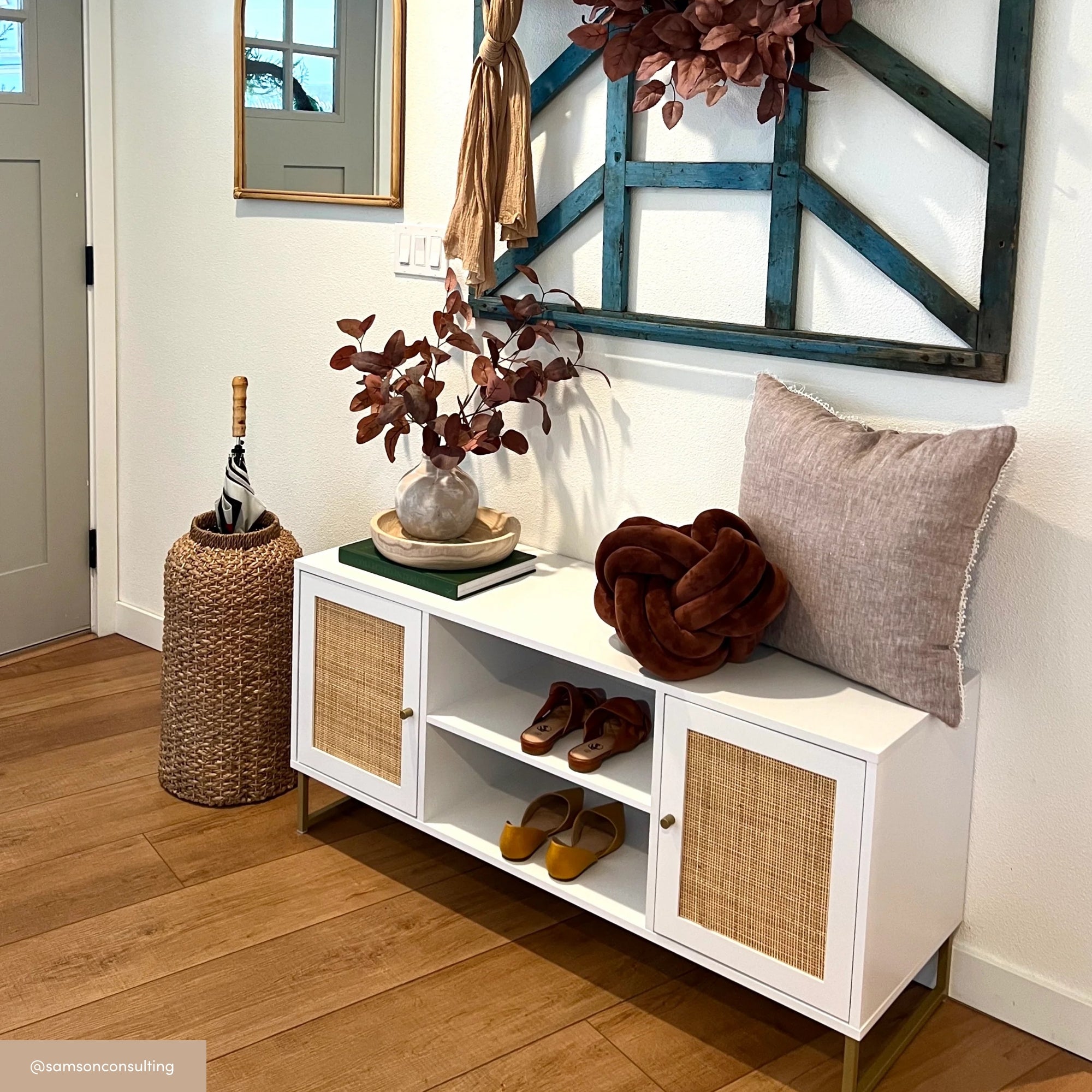 Modern entryway with a white console table, decorative vase with brown foliage, a tan pillow, brown blanket, and shoes. A woven umbrella stand and wall decor with a mirror and wreath are also visible.