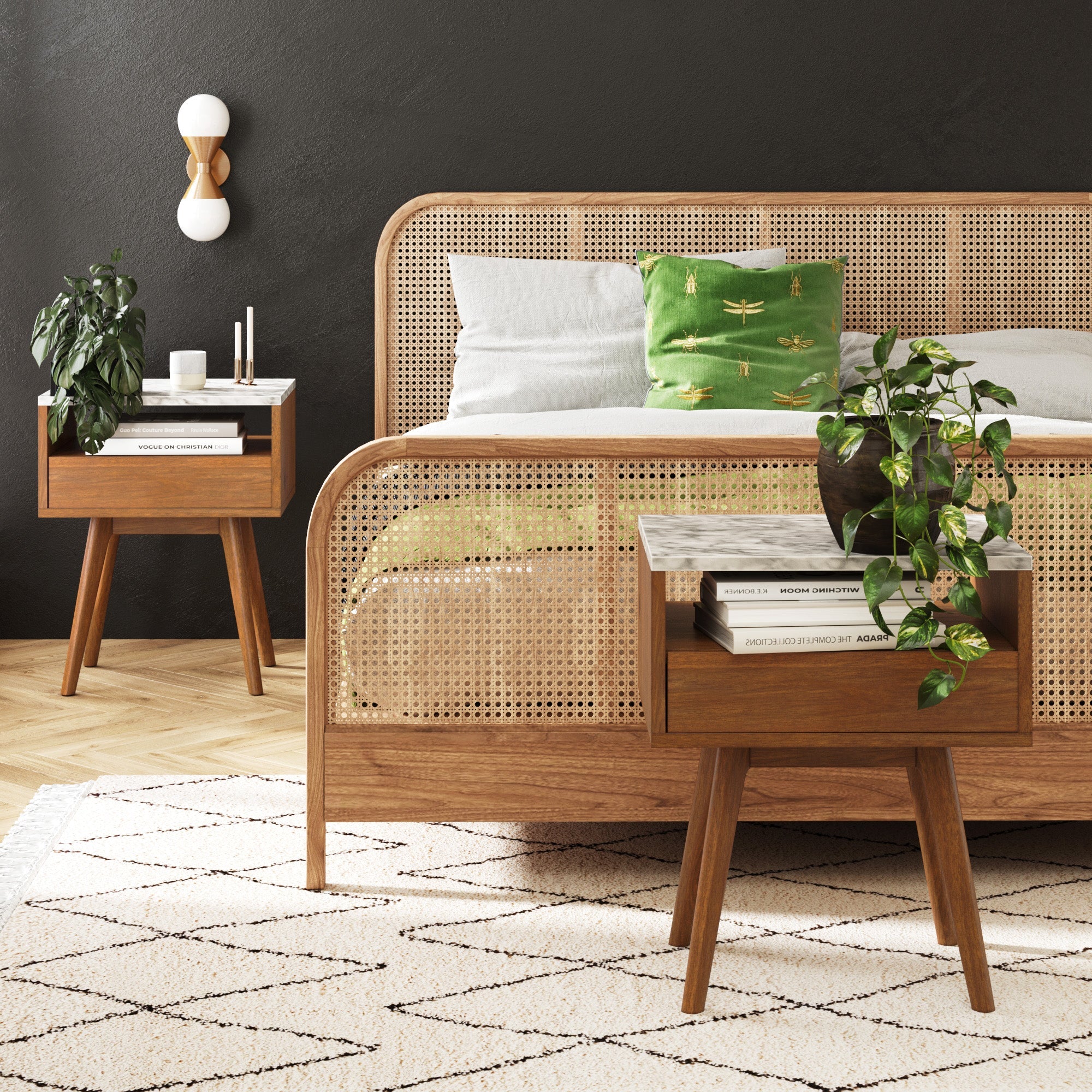 A cozy bedroom with a cane bed frame, green pillow, and two Nathan James Mid-Century Faux Marble Nightstands styled with plants, books, and lamps, set against a dark wall and a light geometric rug on wooden flooring.