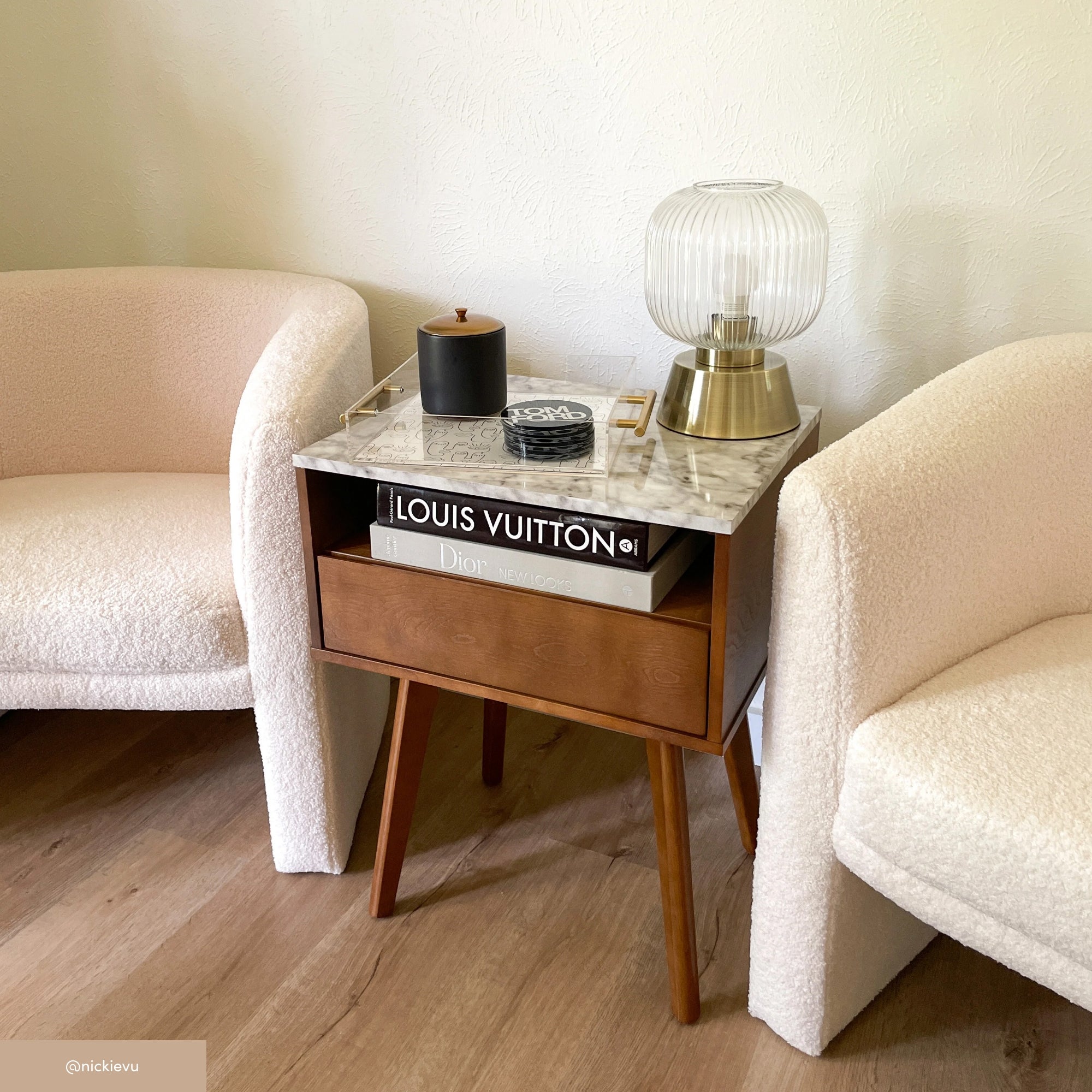 A modern side table with a marble top, books, a candle, and a glass lamp, placed between two cream-colored, curved armchairs on a wooden floor against a light-colored wall.