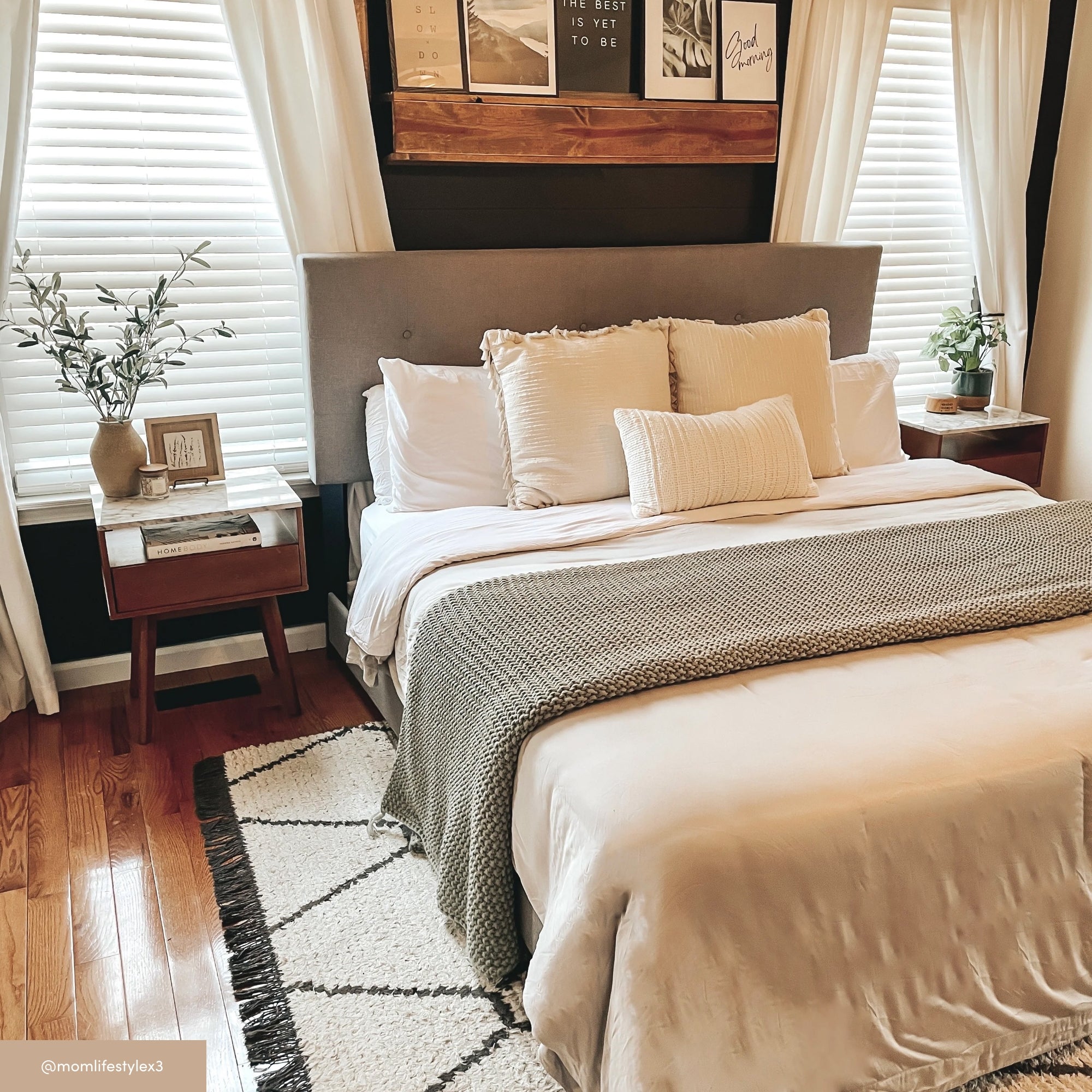 A cozy bedroom with a neatly made bed featuring white and beige bedding, flanked by wooden nightstands and windows with white curtains. Wall art is arranged above the gray headboard, and a soft rug covers the wooden floor.