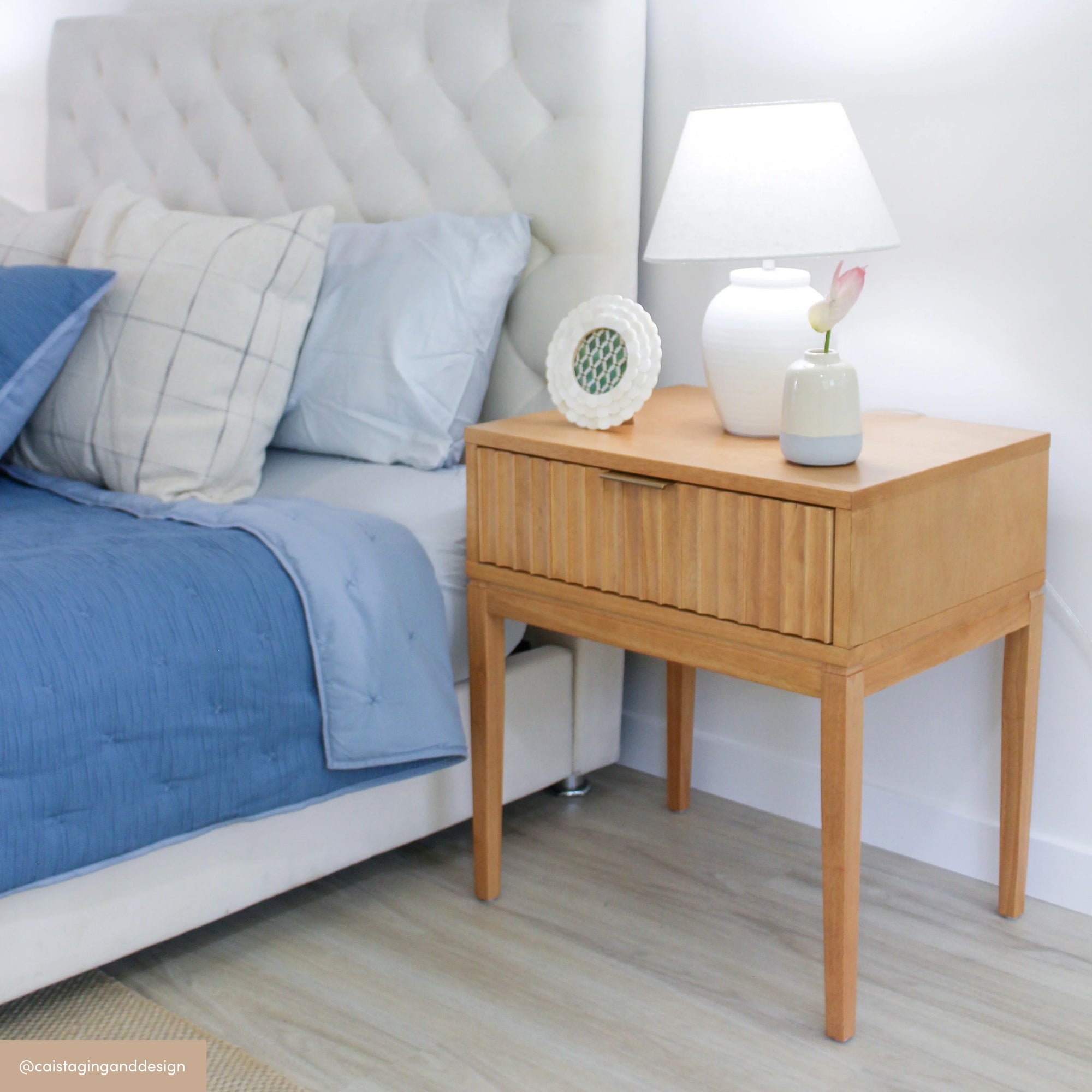 A modern bedroom features a wooden nightstand with a lamp, a small vase with a pink flower, and a decorative plate beside a bed with blue and gray bedding and a tufted white headboard.