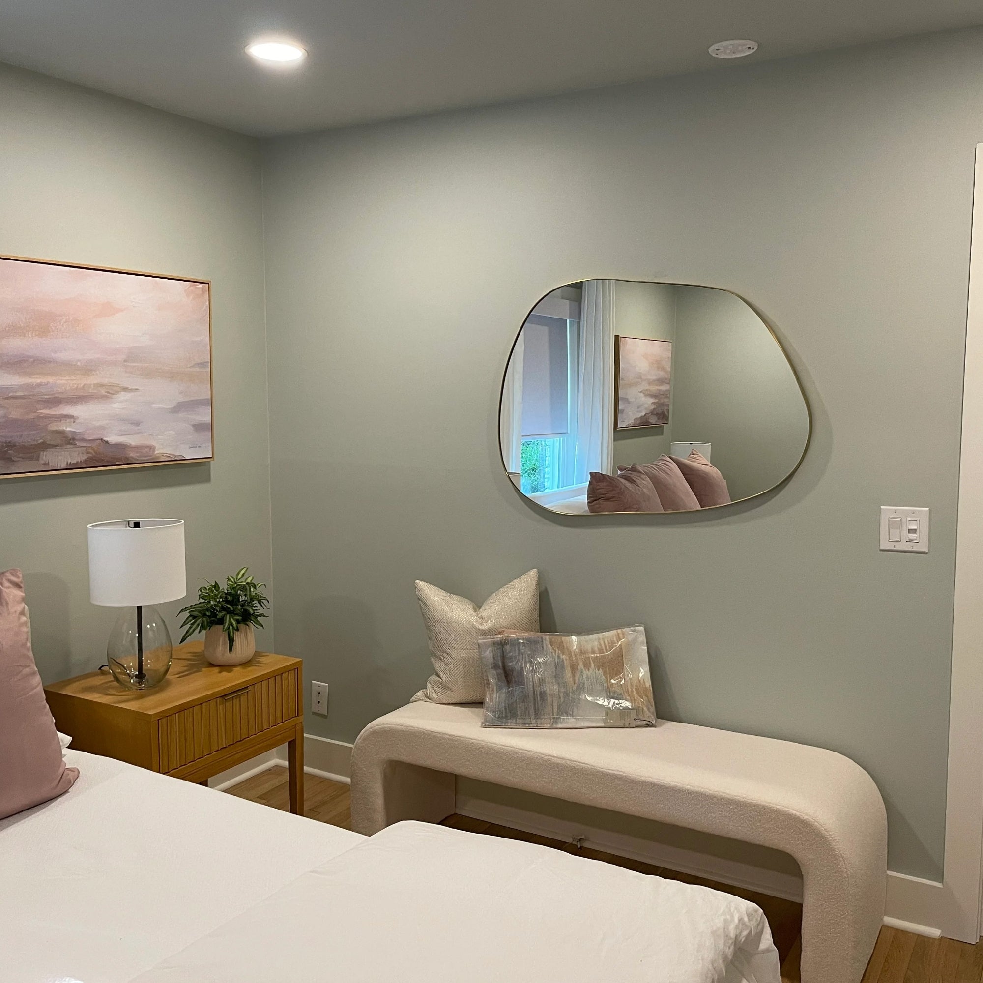 A cozy bedroom with pale green walls, a bed with light pink bedding, a wooden nightstand with a lamp and plant, a modern abstract painting, an irregularly shaped mirror, and a bench with pillows beneath the mirror.