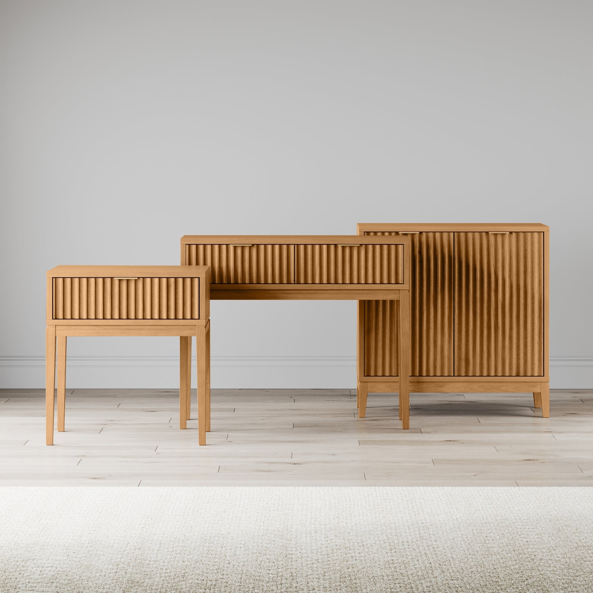 Three wooden furniture pieces with vertical grooves stand on a light wood floor against a plain light gray wall. The set includes a small table, a desk, and a tall cabinet, all with a minimalist design.