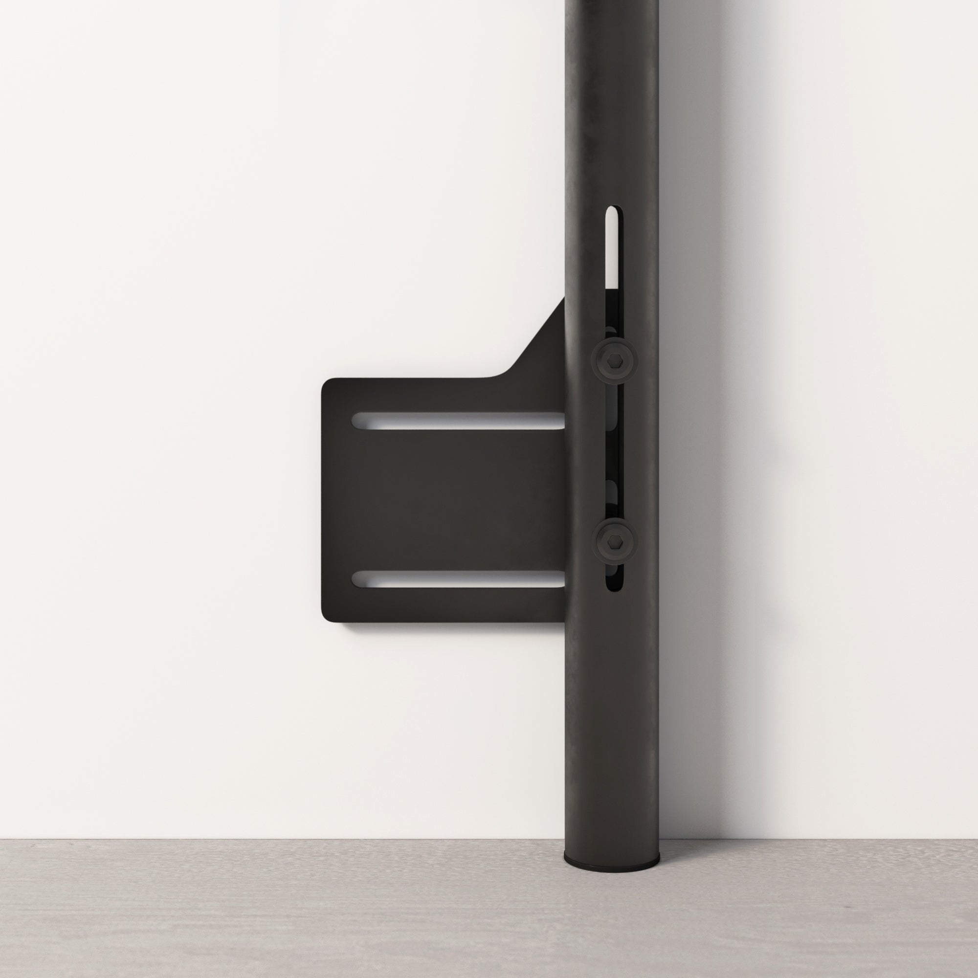A close-up of a sleek black metal bracket attached to a vertical pole, mounted against a white wall with a light gray floor. The bracket has two horizontal slots and visible screw attachments.