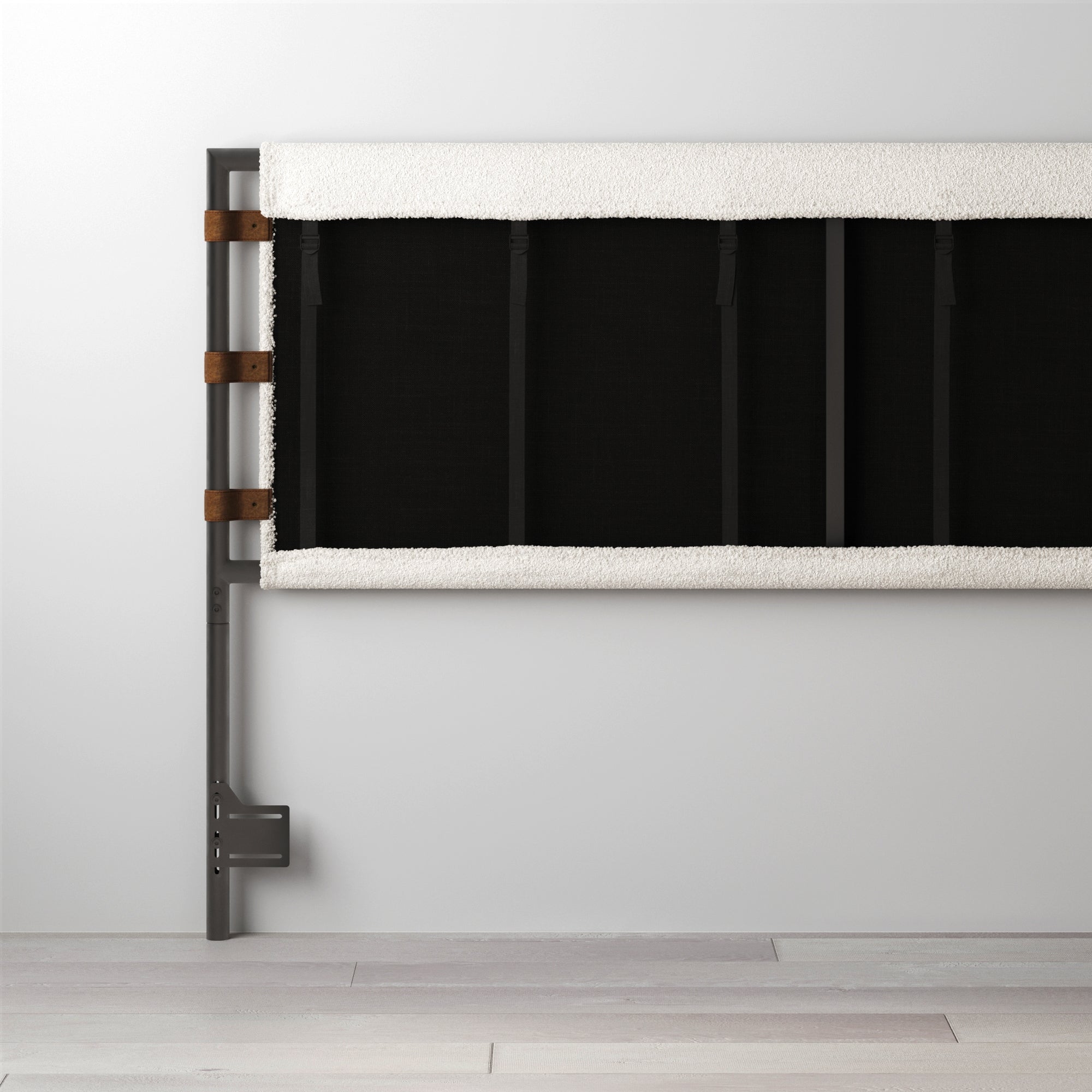 A modern black and white baby crib rail with a metal frame, fabric panel, and brown leather straps, standing against a light gray wall and light wood floor.