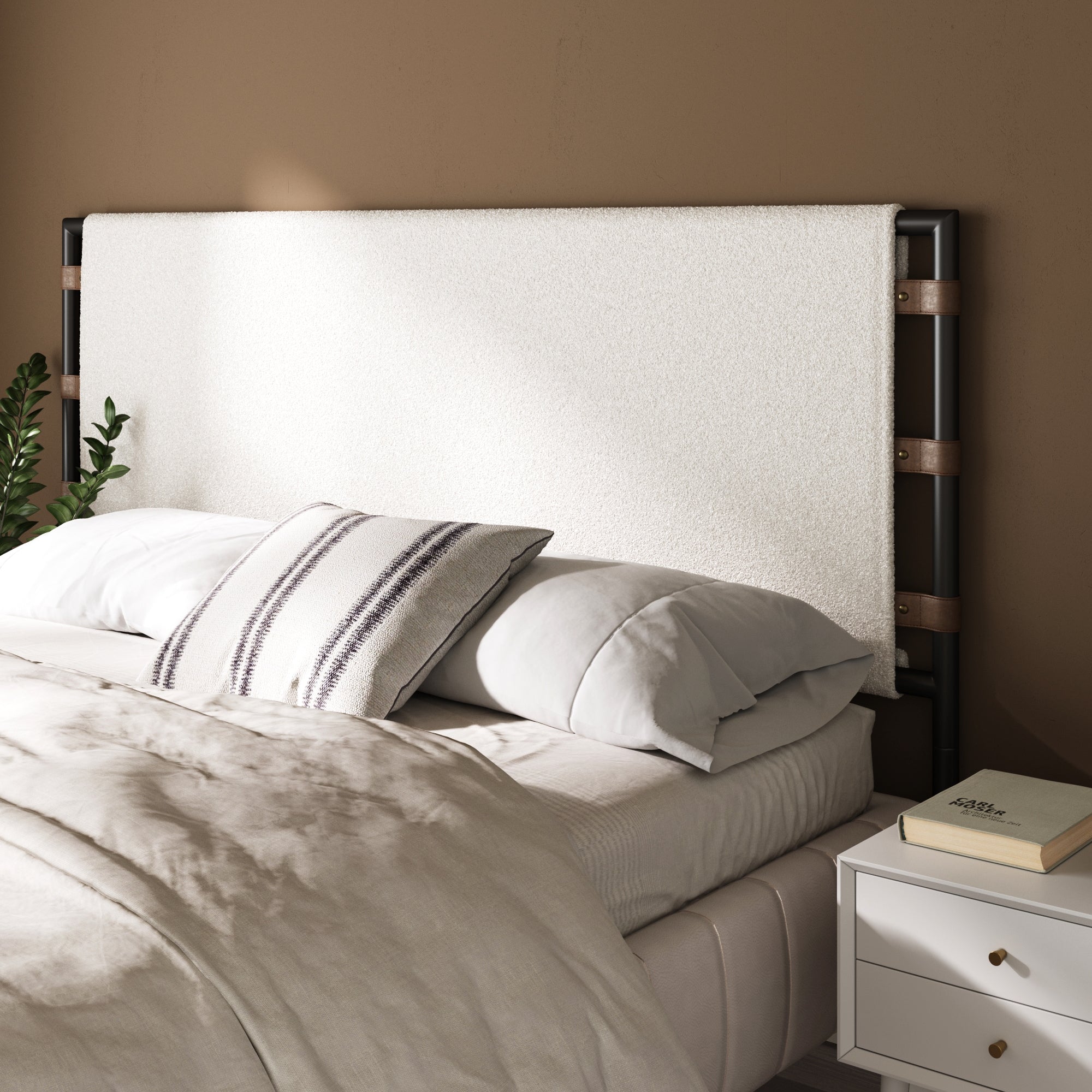 A neatly made bed with light bedding, striped accent pillows, and a white Nathan James Boucle King Upholstered Headboard stands against a beige wall. A white nightstand with a closed book and plant sits beside the bed.