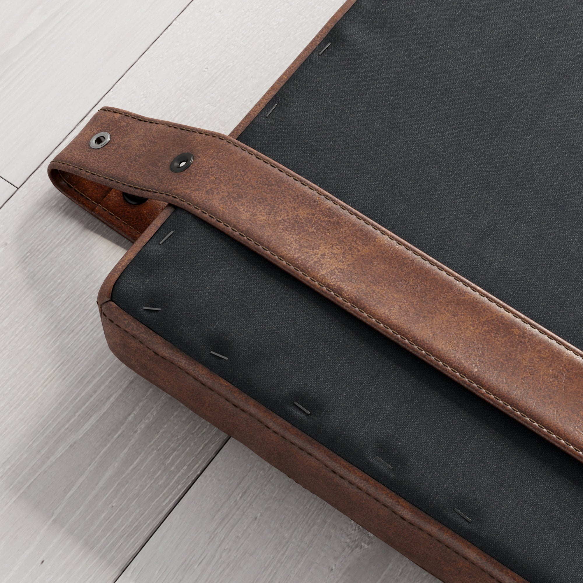 Close-up of a corner of a cushion or upholstered seat with dark fabric on the bottom, brown leather trim, and a brown leather strap attached with a metal rivet, placed on light wood flooring.