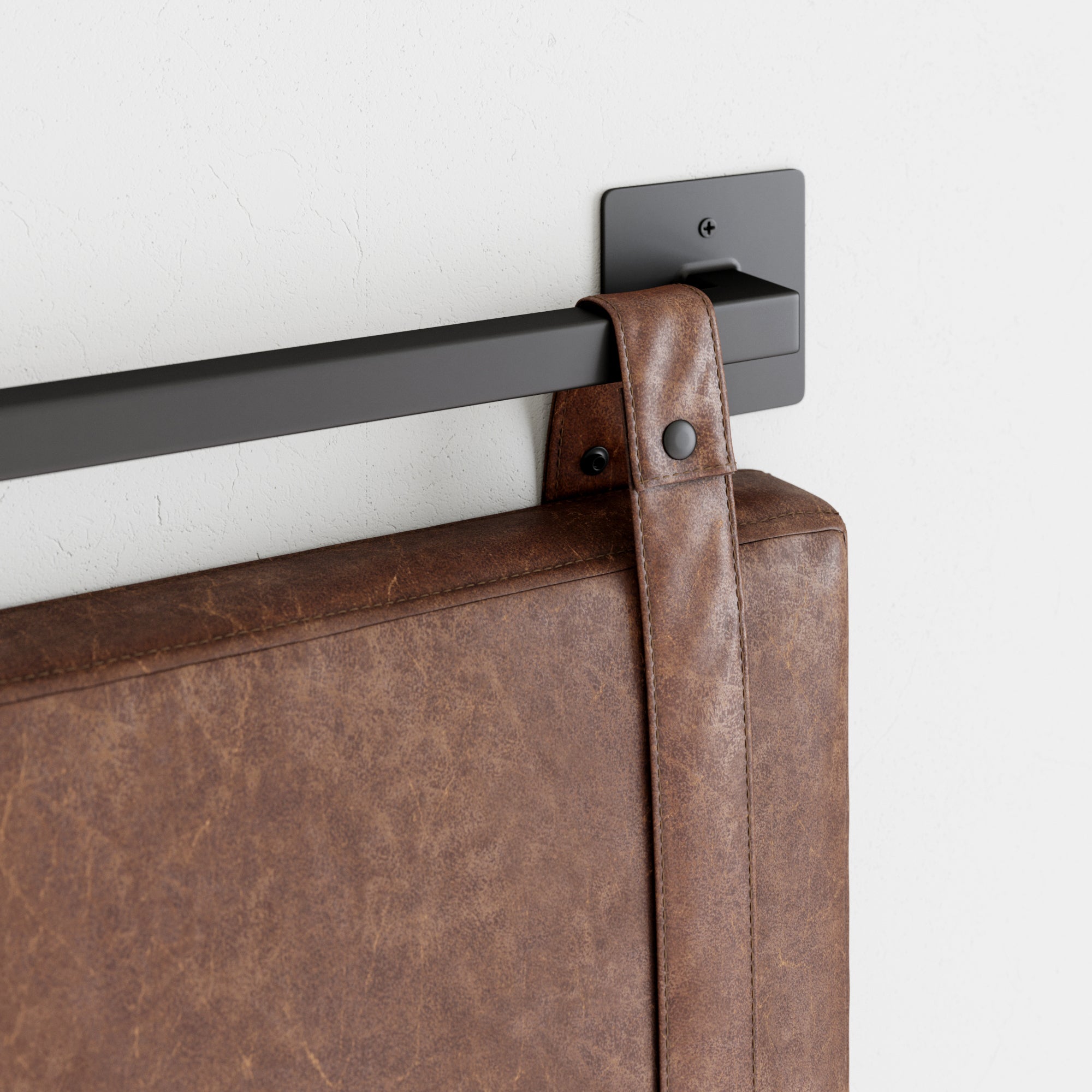 A close-up of a brown leather strap securing a padded headboard to a black metal wall-mounted bracket against a white textured wall.