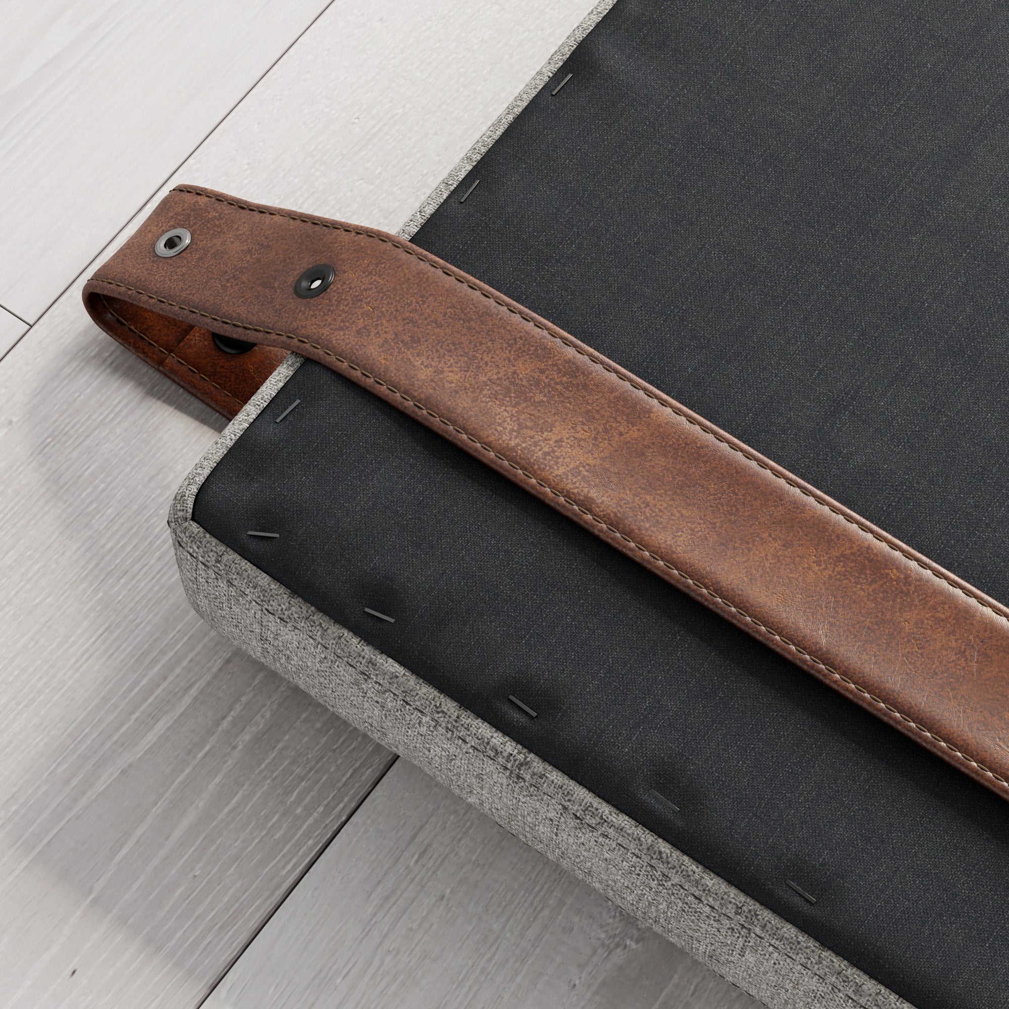 A close-up of a brown leather strap attached to the corner of a gray upholstered cushion or mattress, placed on a light wood floor. The strap is secured with metal rivets.