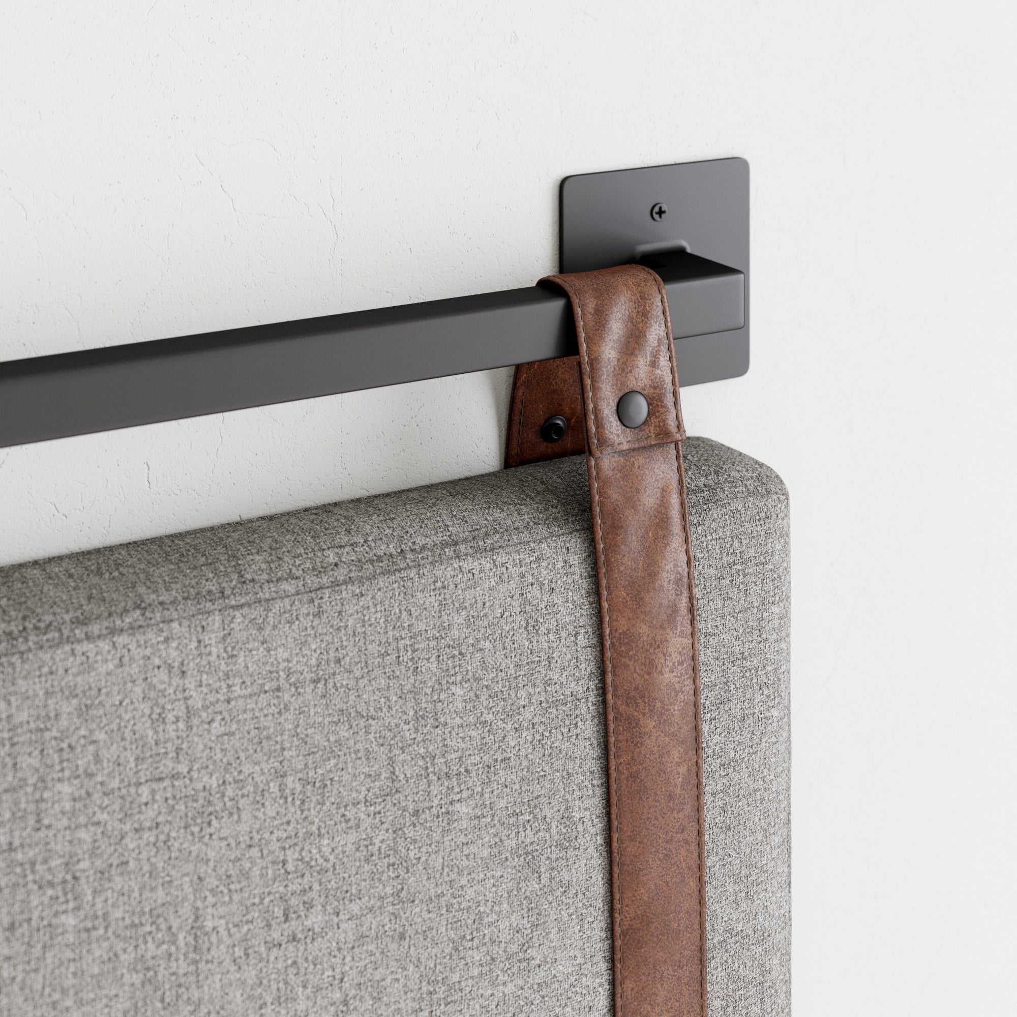 Close-up of a gray upholstered headboard secured to a black metal wall-mount bar with a brown leather strap and black hardware against a white wall.