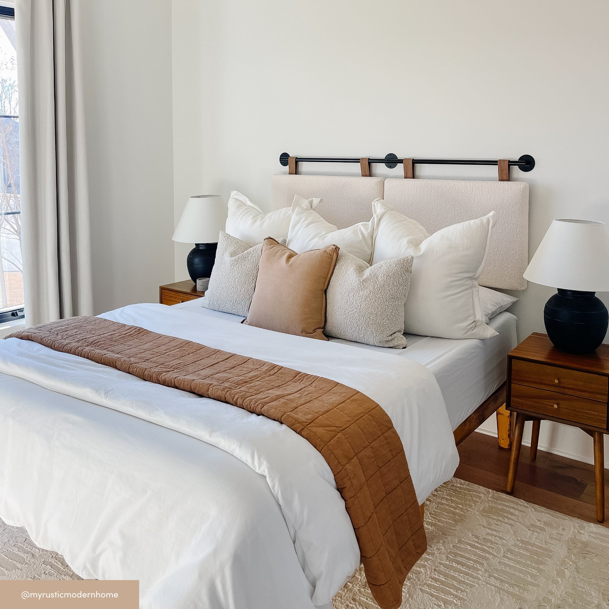 A neatly made bed with white bedding, brown throw blanket, and multiple pillows sits between two wooden nightstands with black lamps, in a bright, modern bedroom with cream walls and a large window.