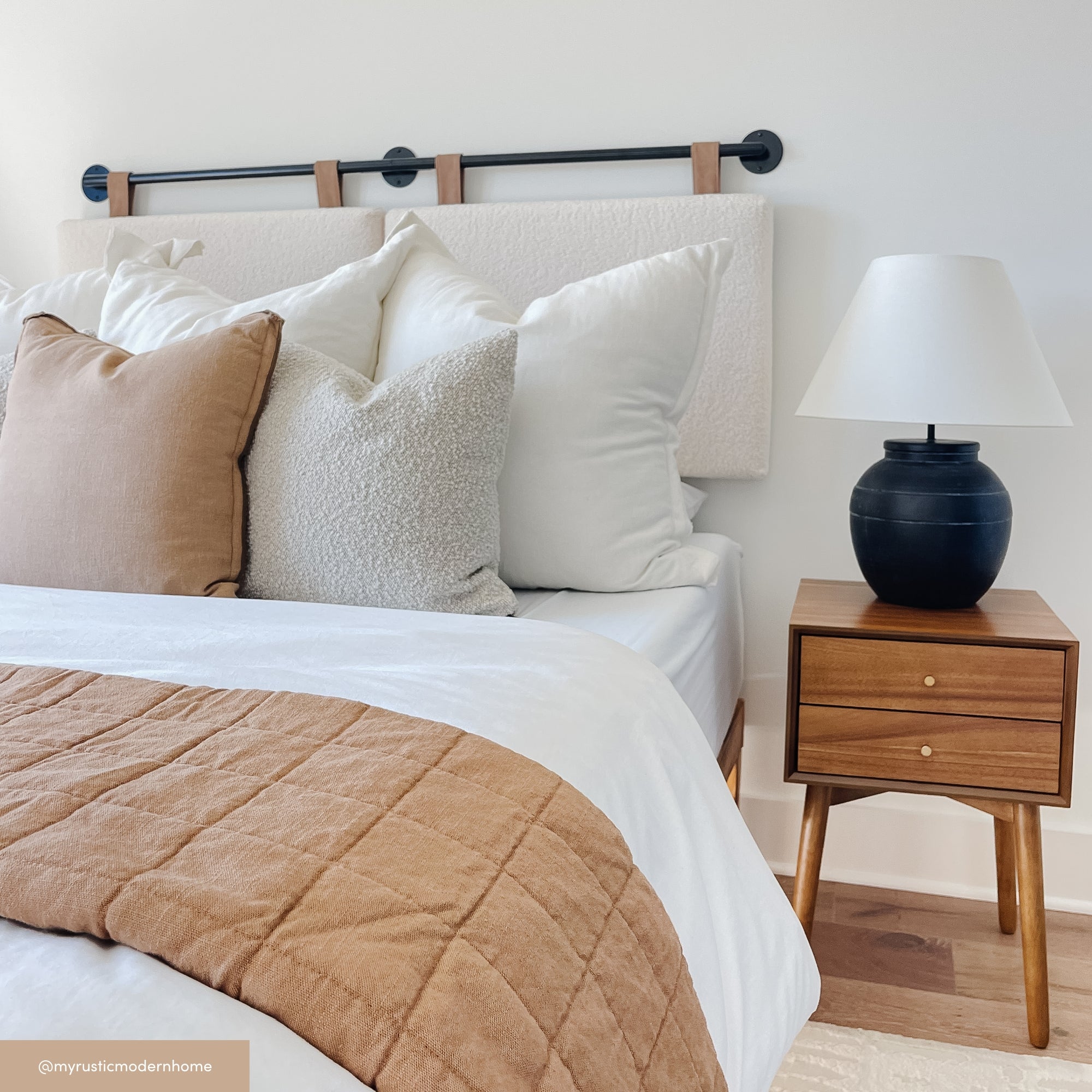 A neatly made bed with white and beige pillows, a tan quilt, and a wooden nightstand holding a black lamp with a white shade, in a modern, minimalist bedroom.