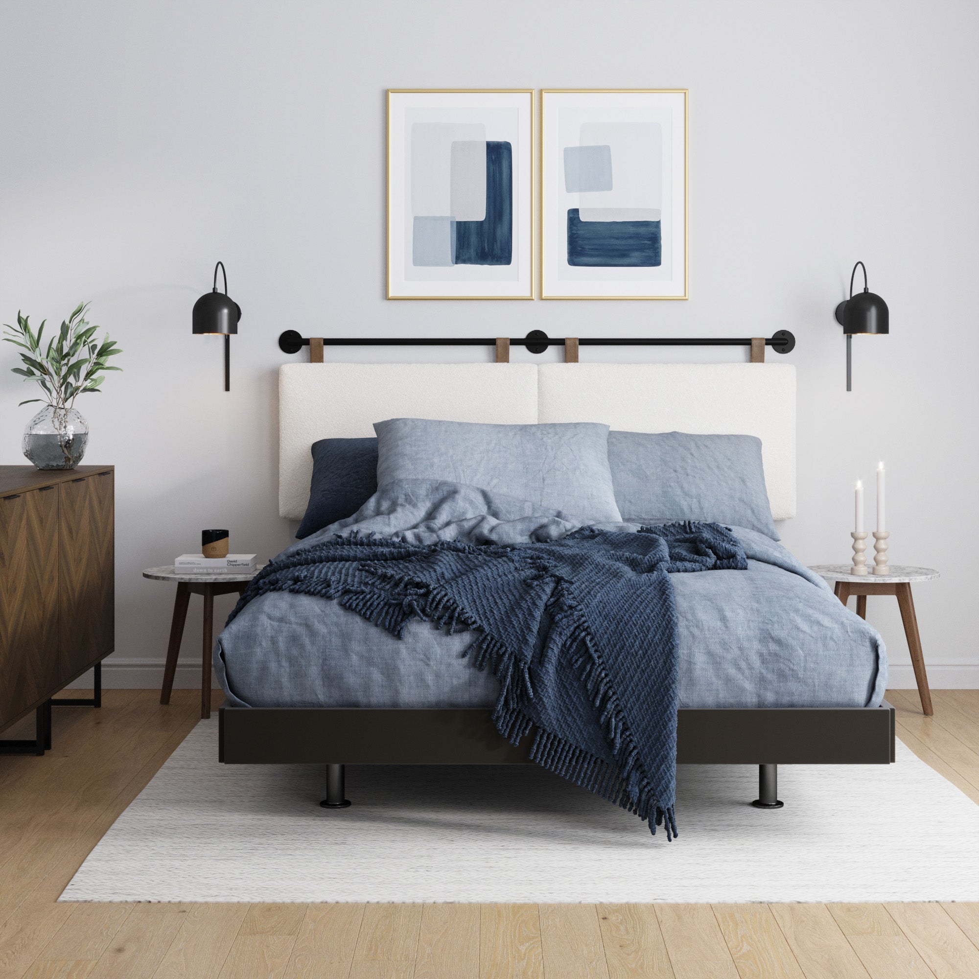 A modern bedroom with a neatly made bed featuring blue and gray bedding, matching throw blanket, two abstract framed prints above the headboard, wall-mounted lamps, side tables, and a dresser with a plant.