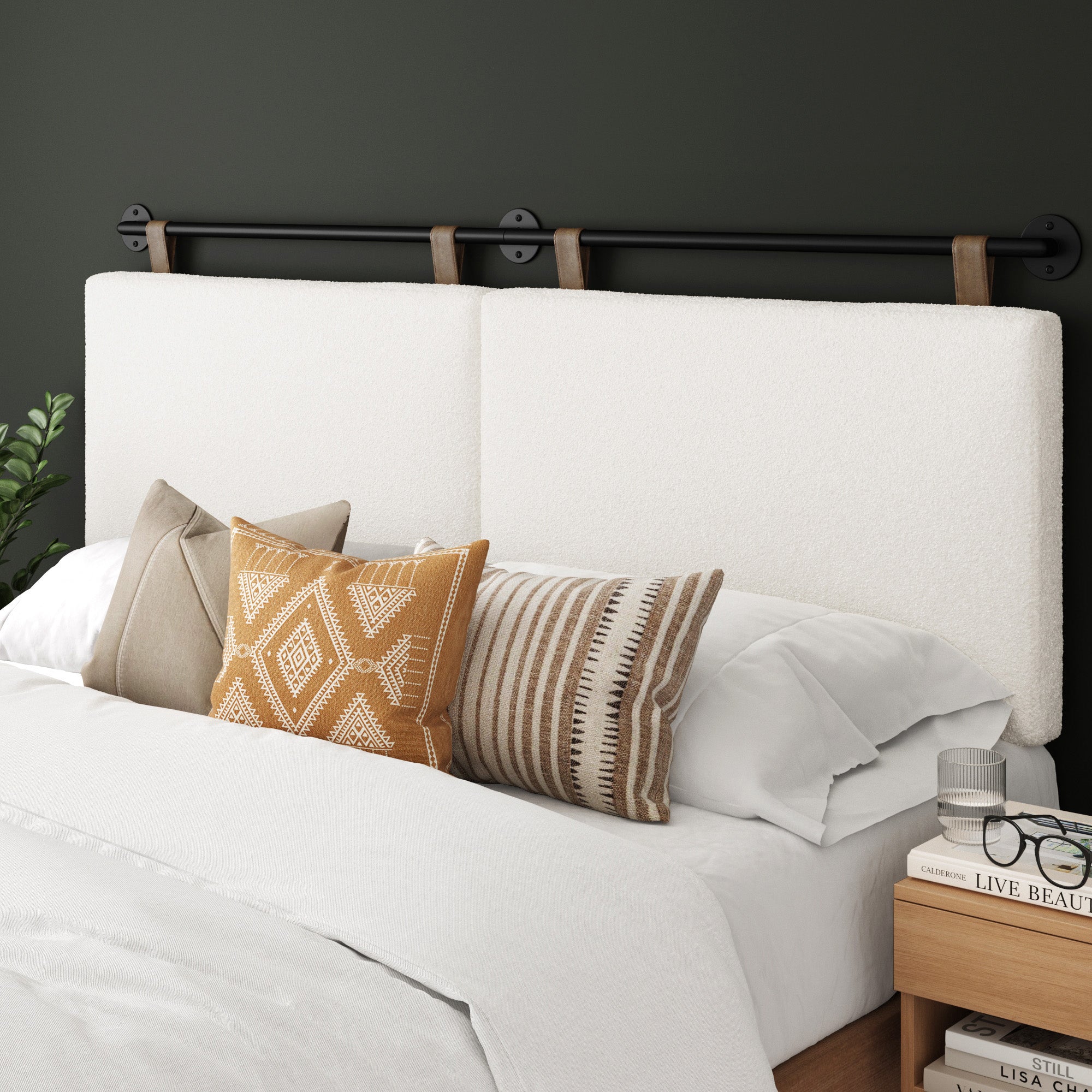 A modern bed with a white, fabric-upholstered headboard, decorated with various patterned throw pillows in beige and brown tones. A small plant and stacked books sit on the wooden nightstand beside the bed.