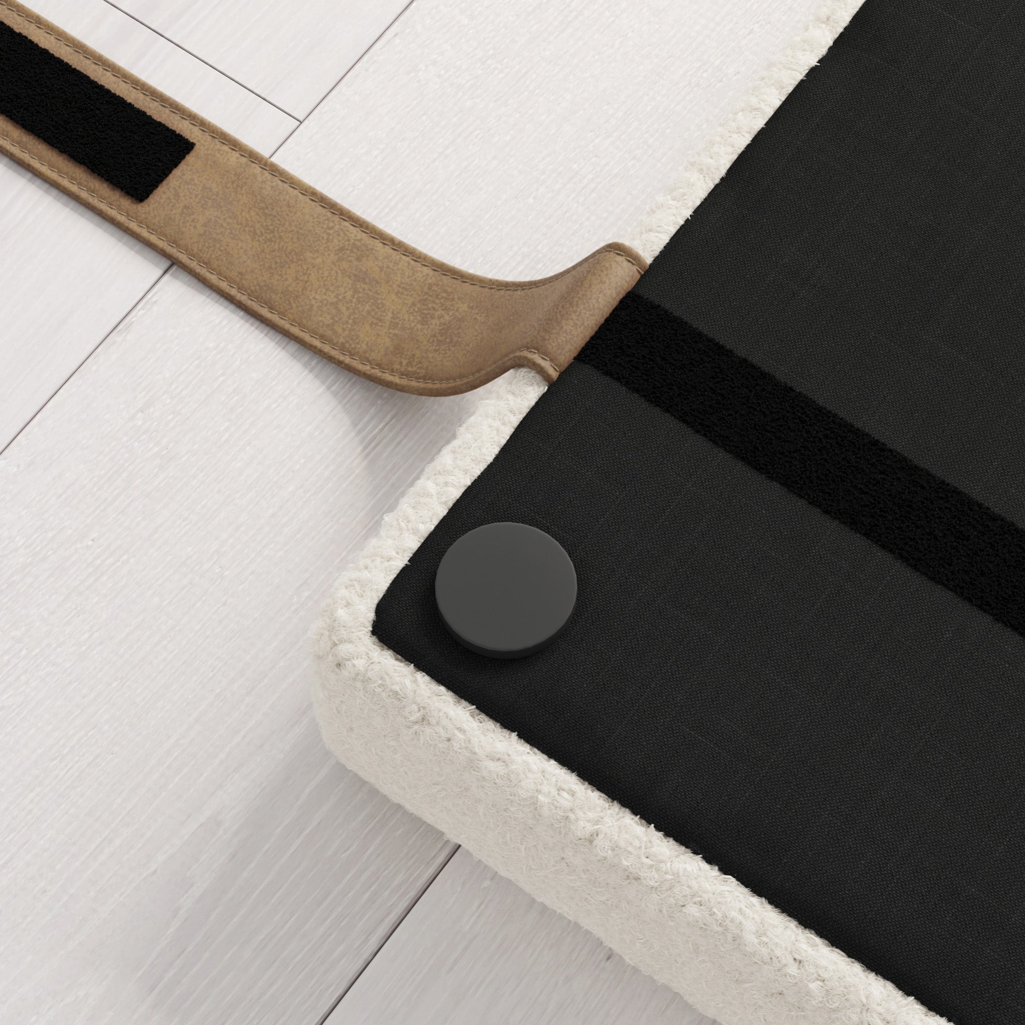 A close-up of a cushioned bench with a black fabric top, a round black button, and a tan leather strap on a light wood floor.